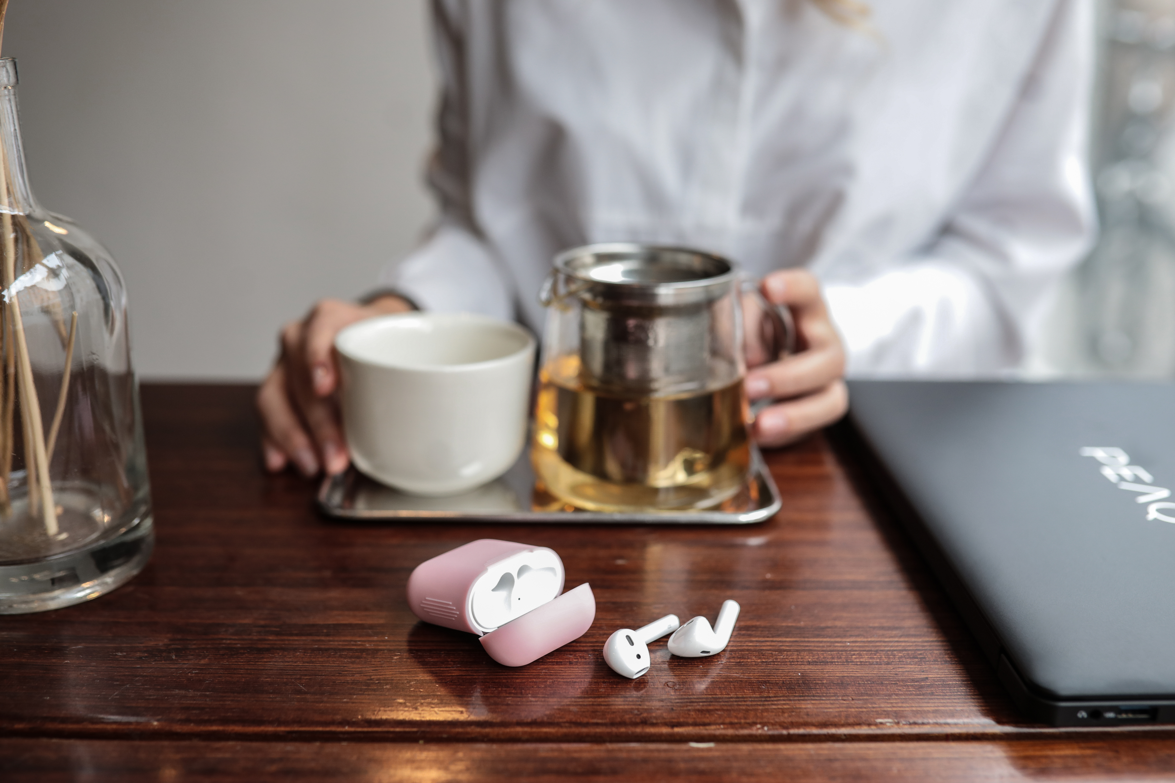 Person hält Tasse und Glasteekanne. AirPods und Etui auf dem Tisch. Laptop im Hintergrund.