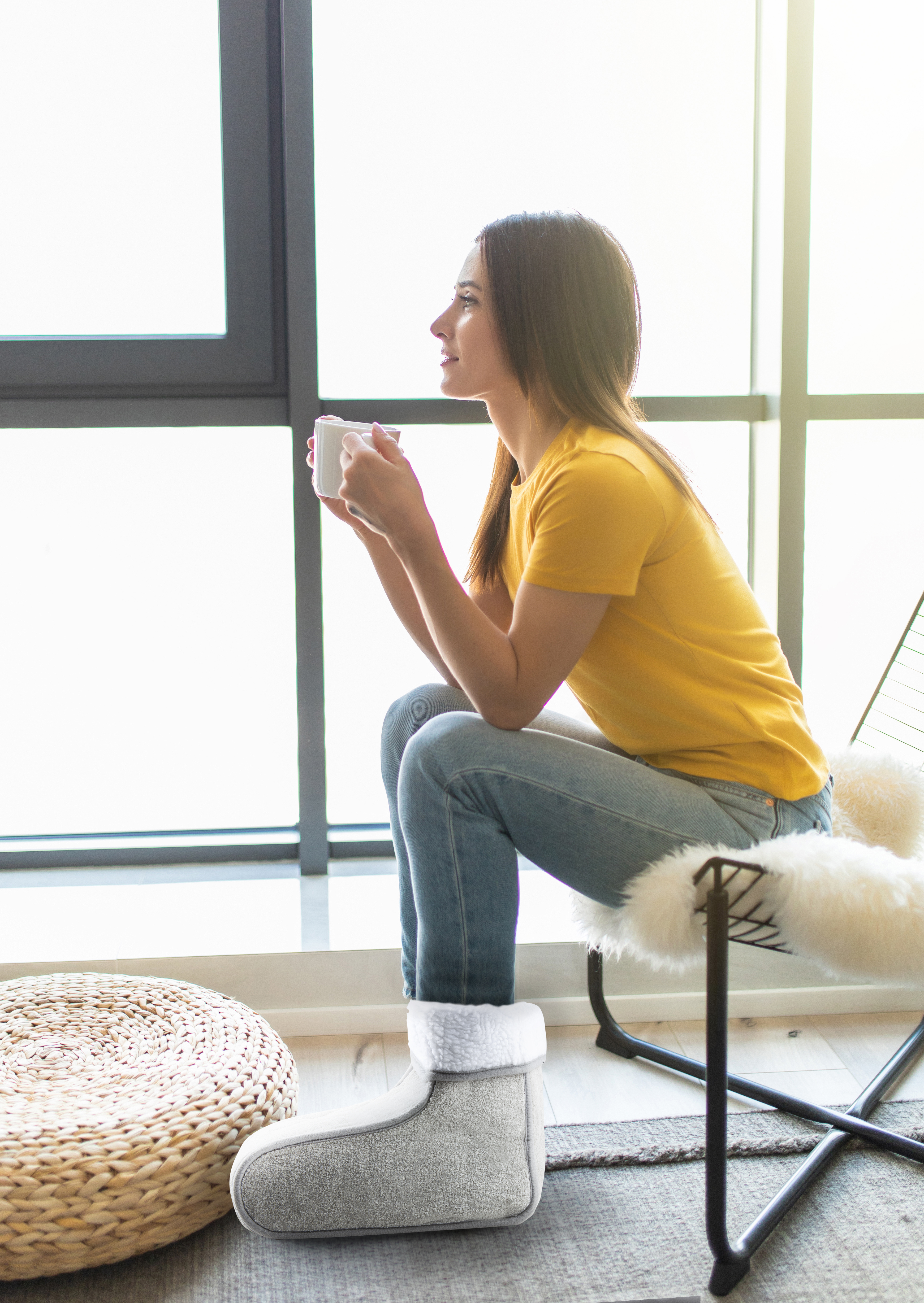 Woman in yellow shirt and jeans, holding a mug, sitting by a window in a chair with a footrest.