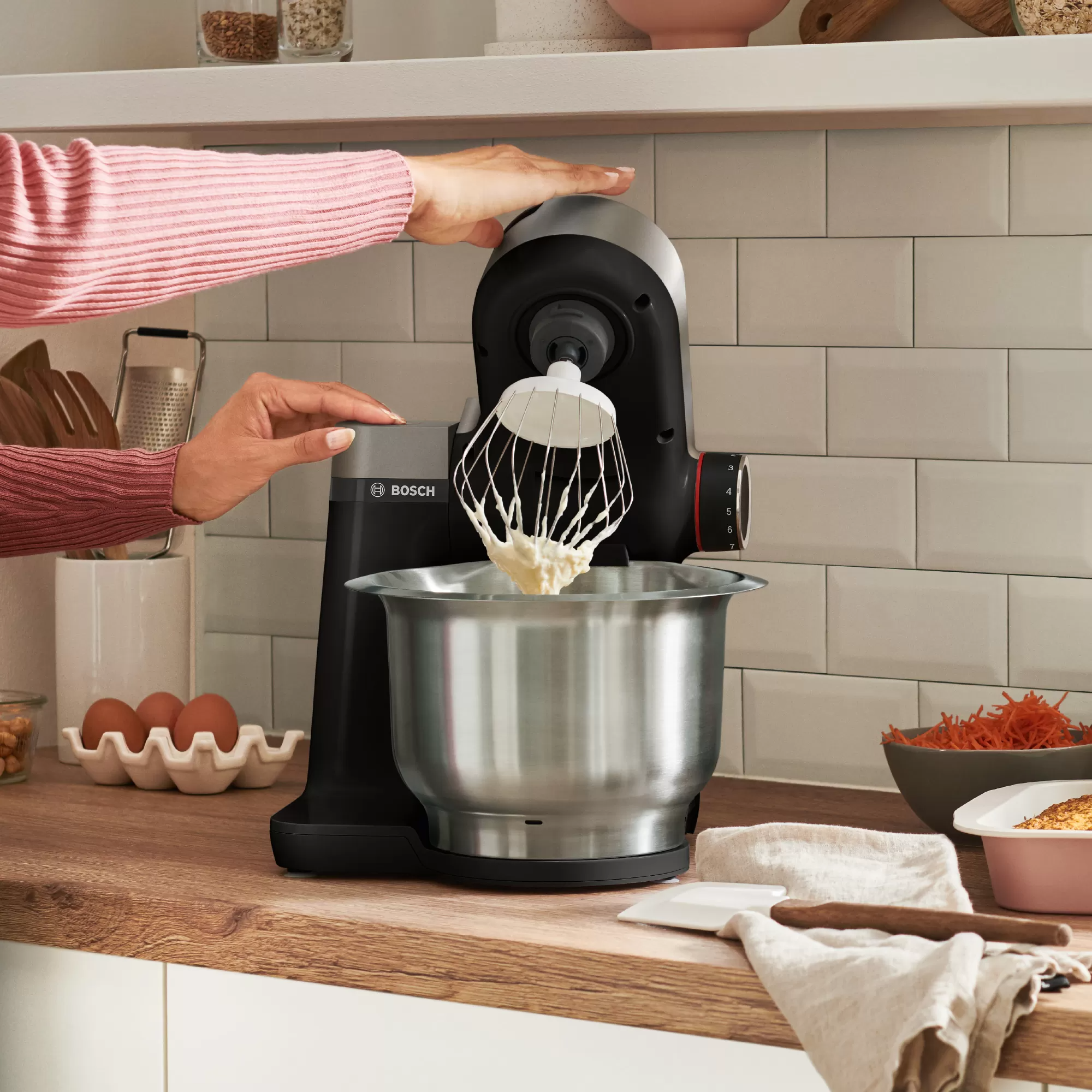 Een vrouw gebruikt een Bosch keukenmachine. Het apparaat is zwart, met een zilveren kom.