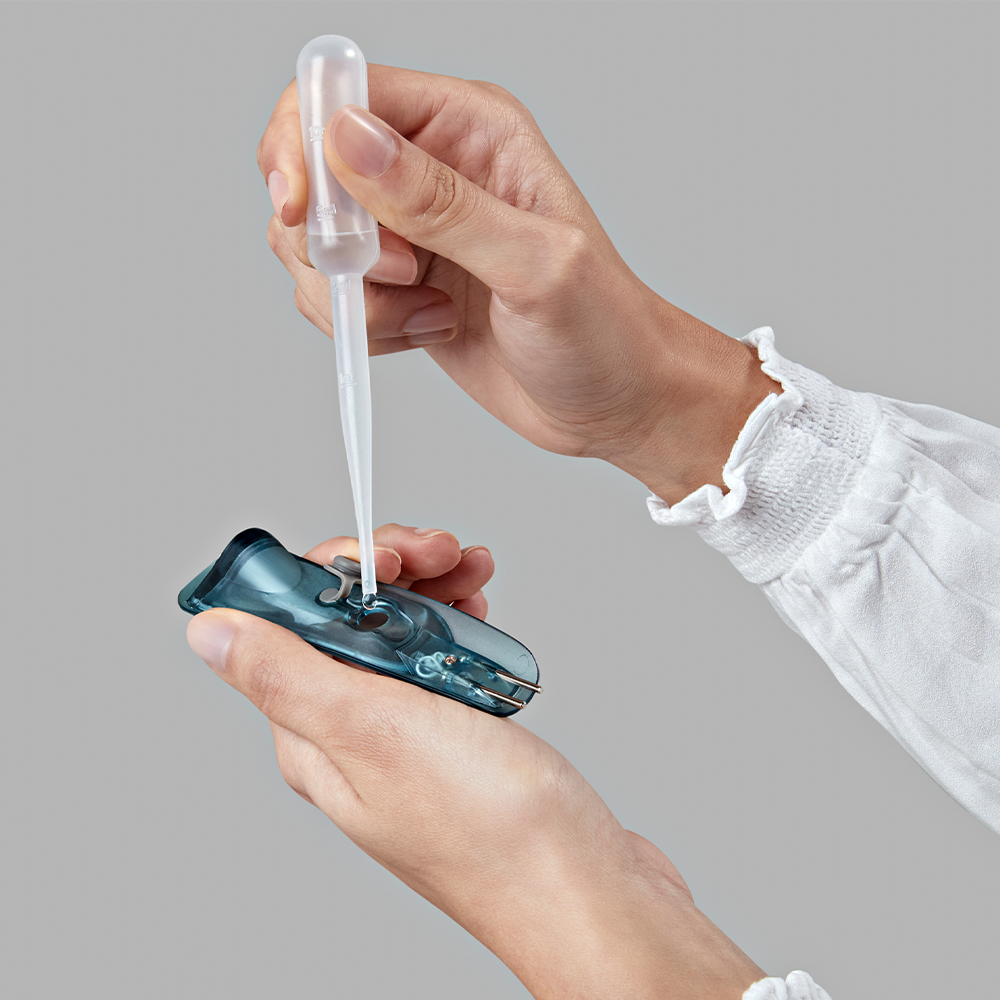 Hands holding a pipette over a blue, open container. Light gray background.