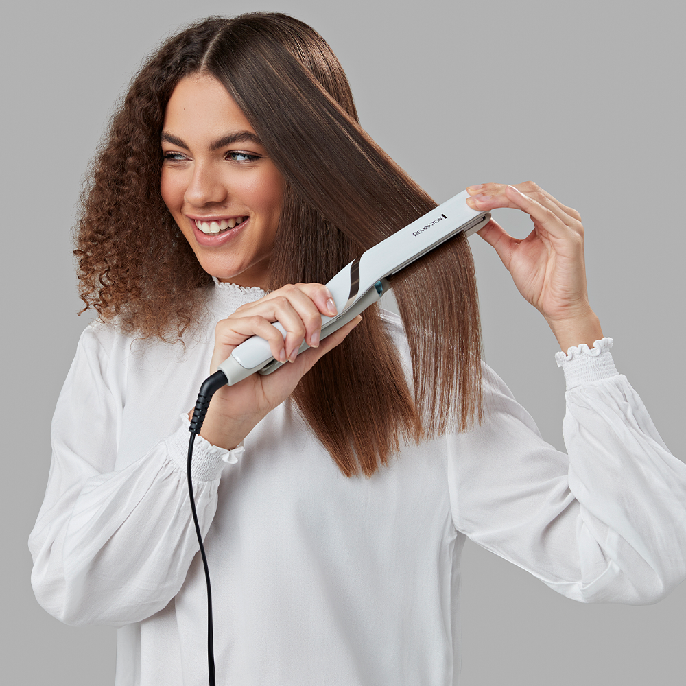 Woman with long hair straightens her hair with a flat iron, smiling at the viewer.