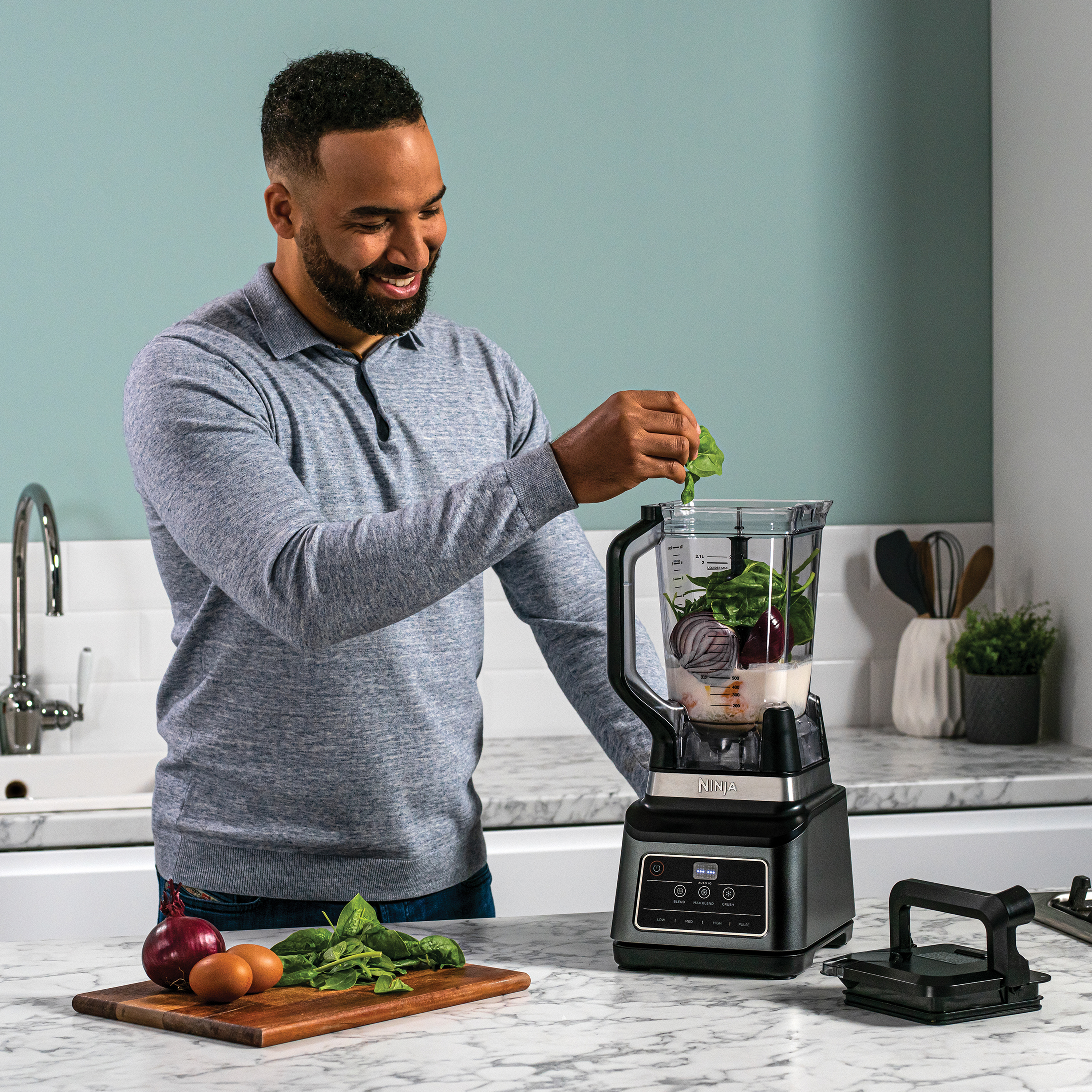 A man adds spinach to a blender with ingredients on a wooden board nearby. Kitchen setting.