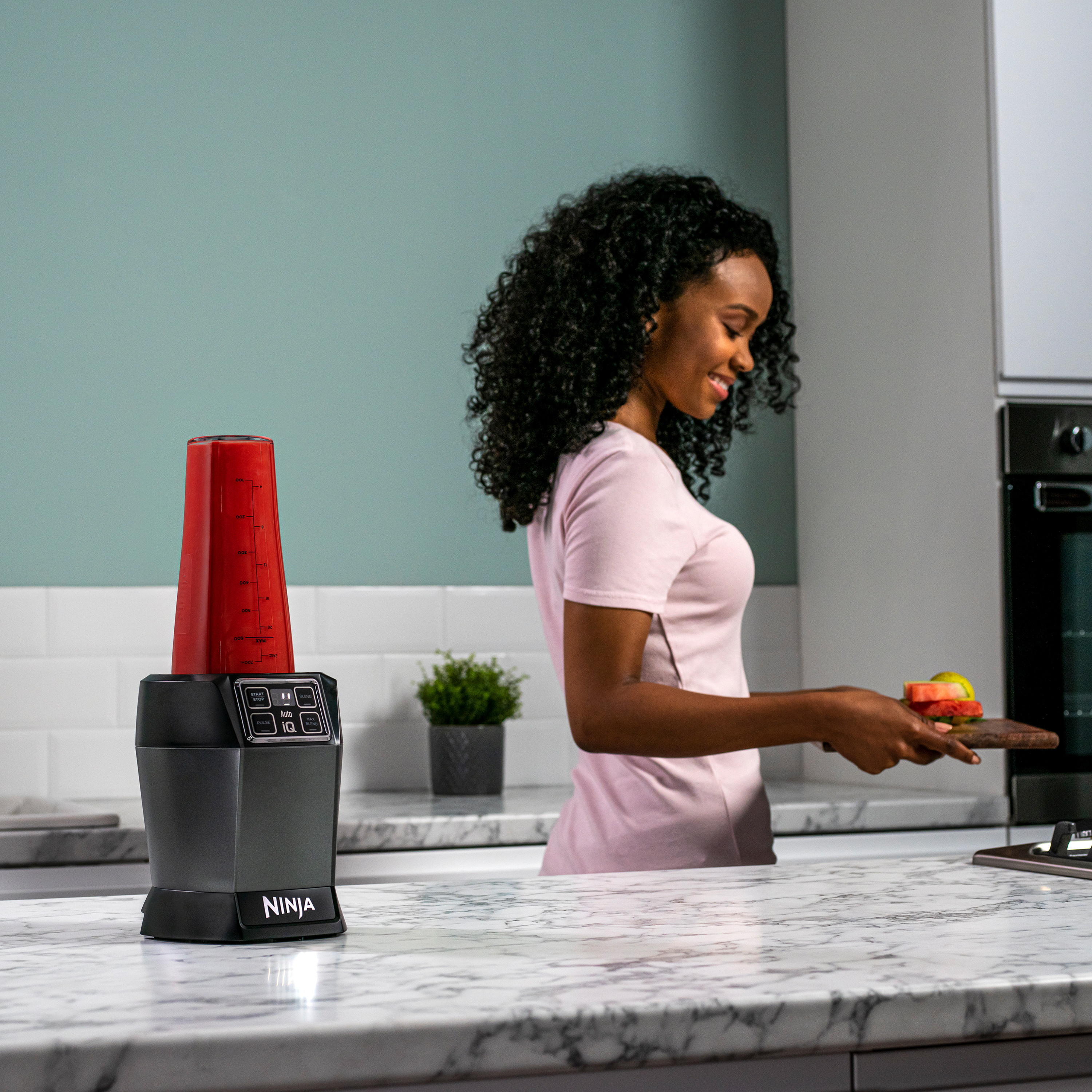 Une femme préparant des aliments avec un mixeur rouge sur un comptoir, souriant.