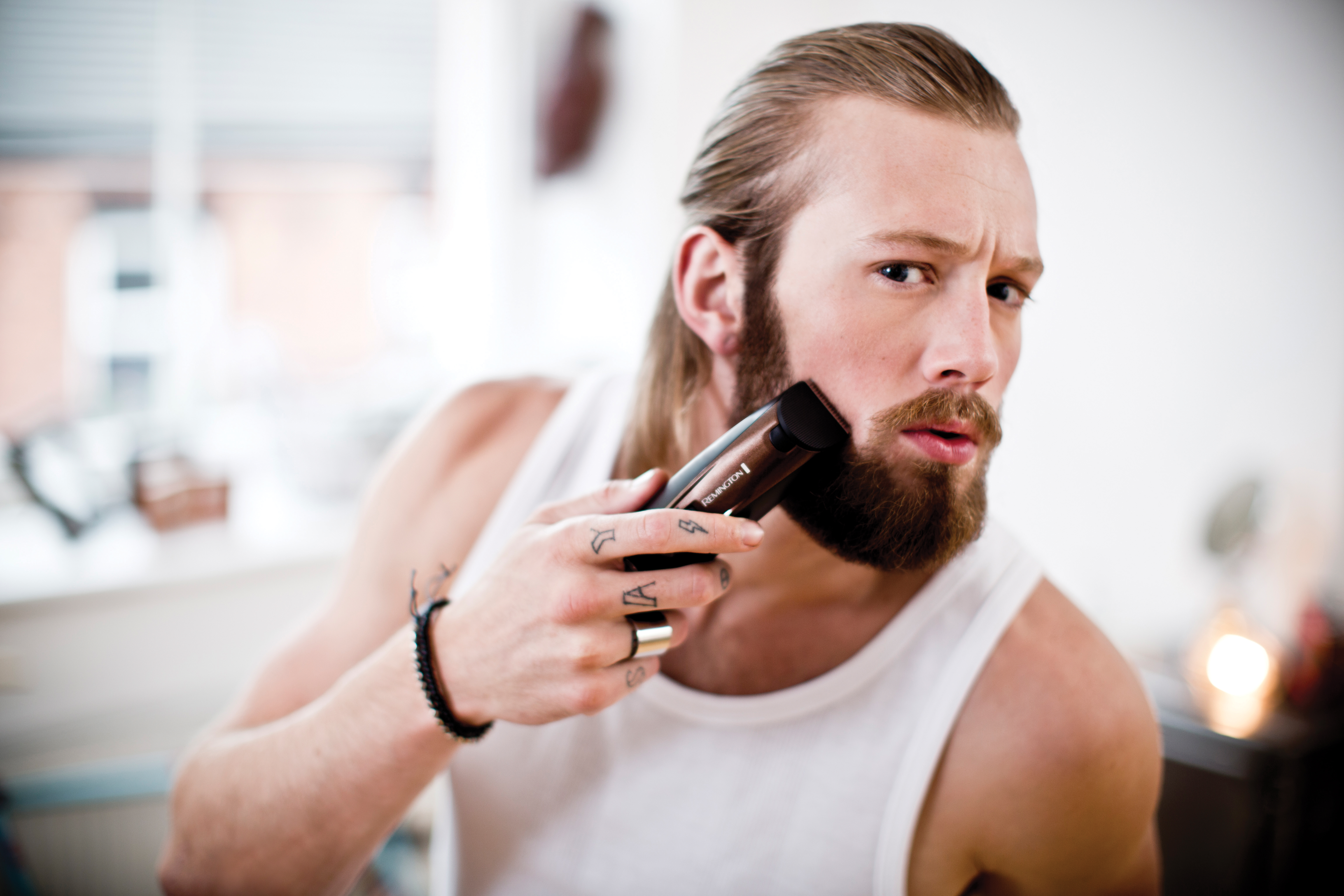 Man shaving beard with a trimmer. He is in a white tank top and has tattoos.