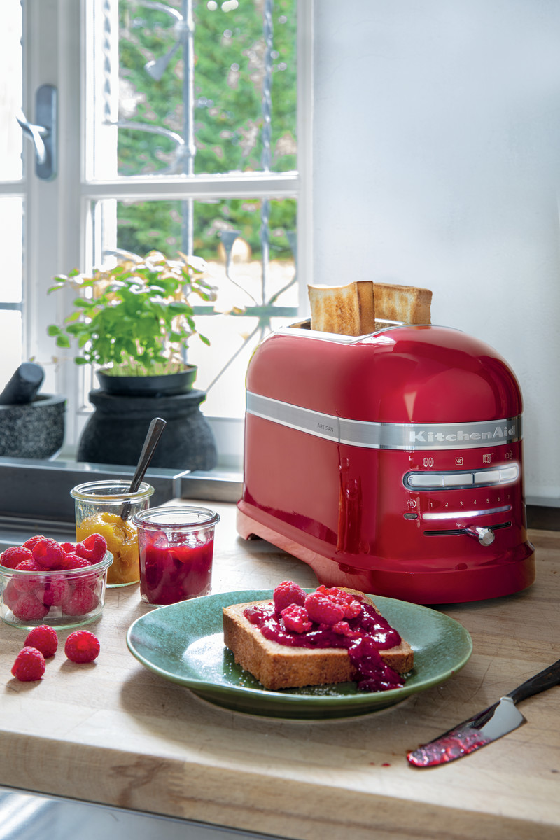 Ein roter KitchenAid-Toaster mit Toast, Marmelade und Himbeeren auf einer Holztheke.