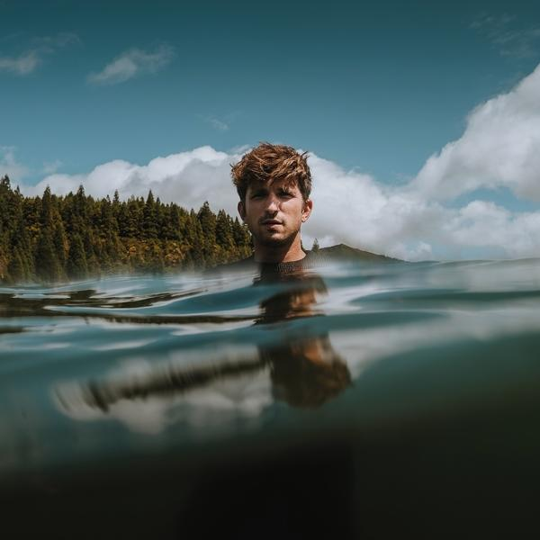 Homme dans l'eau jusqu'au cou, fond forestier, ciel bleu et nuages.