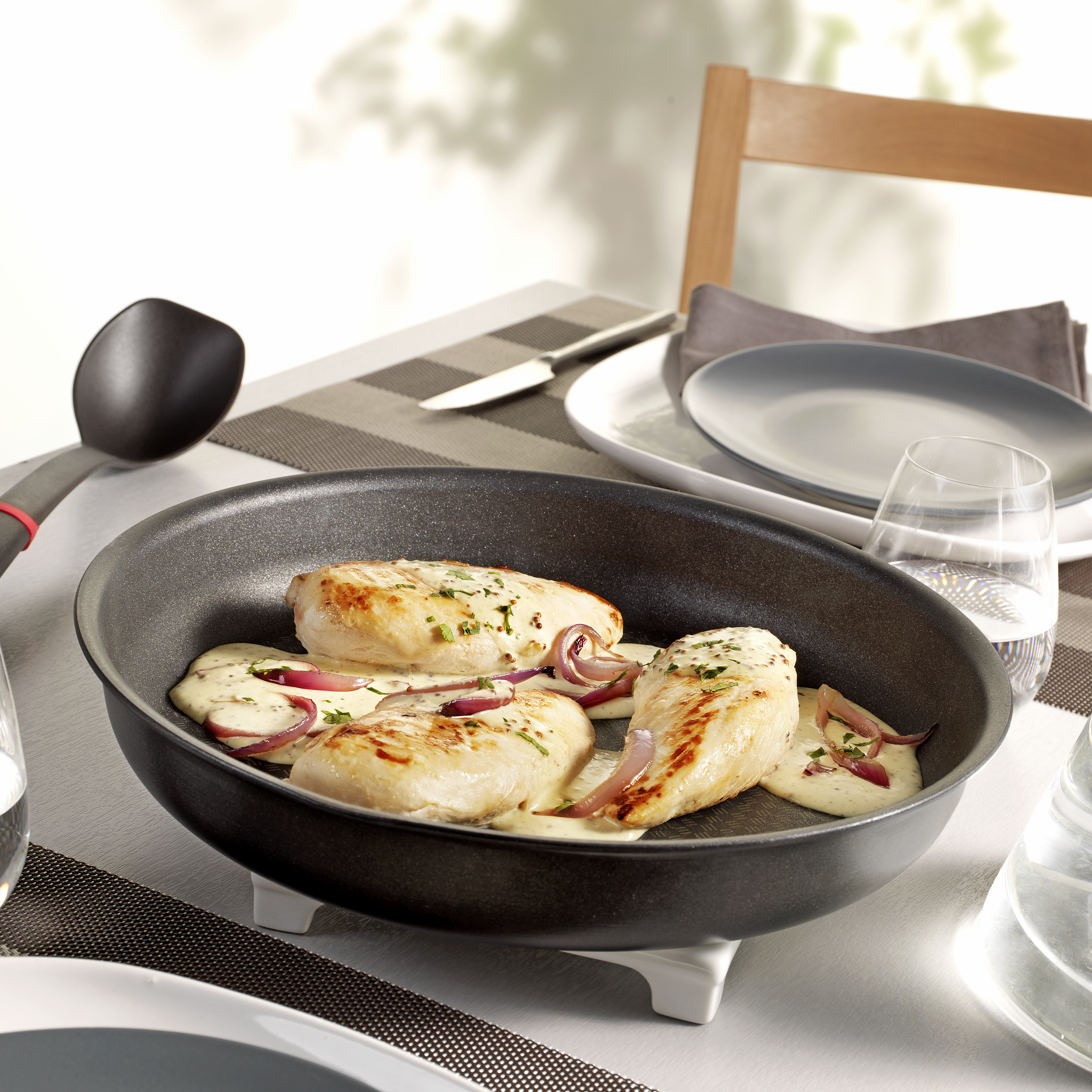 Poitrines de poulet dans une poêle avec sauce et oignons. Table avec assiettes, verre et chaise en arrière-plan.