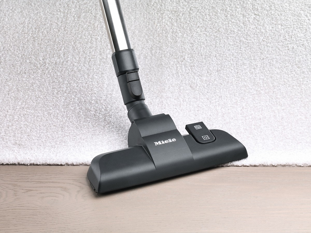 Testa di aspirapolvere nera su pavimento in legno, con tappeto bianco sullo sfondo.