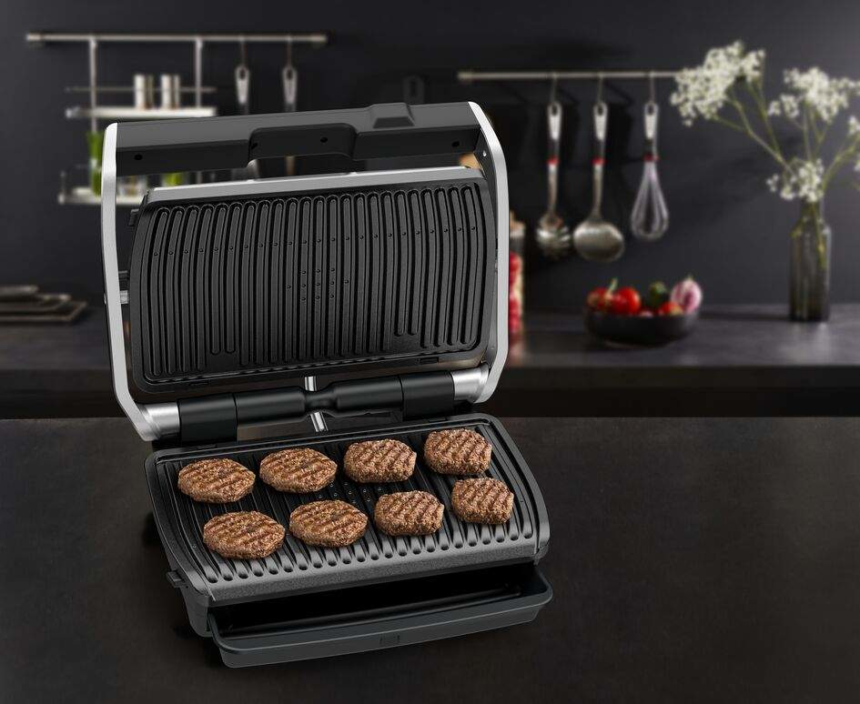 Egy grill, melynek felületén 7 hamburgerpogácsa sül. A grill nyitva van, és fekete felületen.