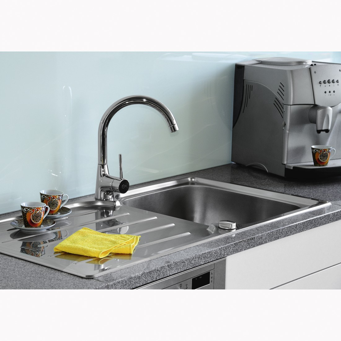 Küchenszene mit silberner Spüle, Wasserhahn und Kaffeemaschine. Zwei Tassen stehen auf einer Theke mit gelbem Tuch.