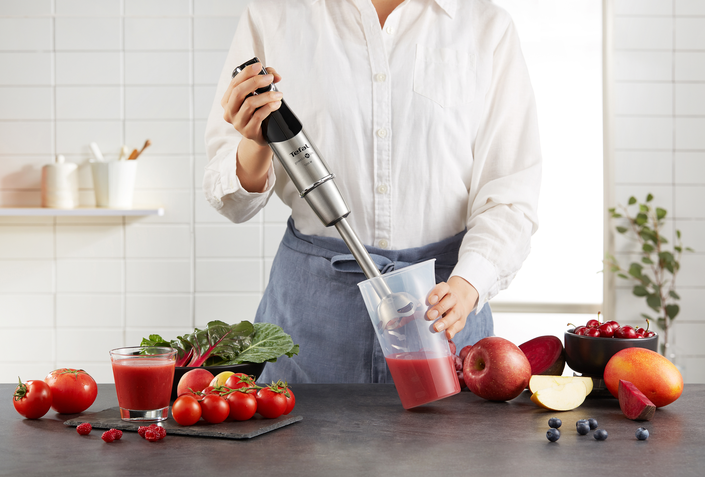Egy személy Tefal botmixert használ. A hozzávalók az asztalon: paradicsom, alma stb.