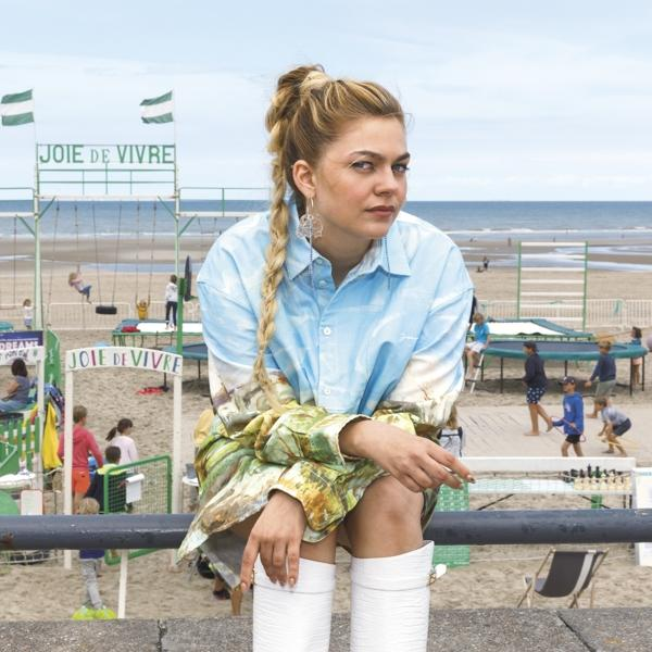 Vrouw zittend op een muur op het strand. Tekst: Joie de Vivre op vlaggen en borden.