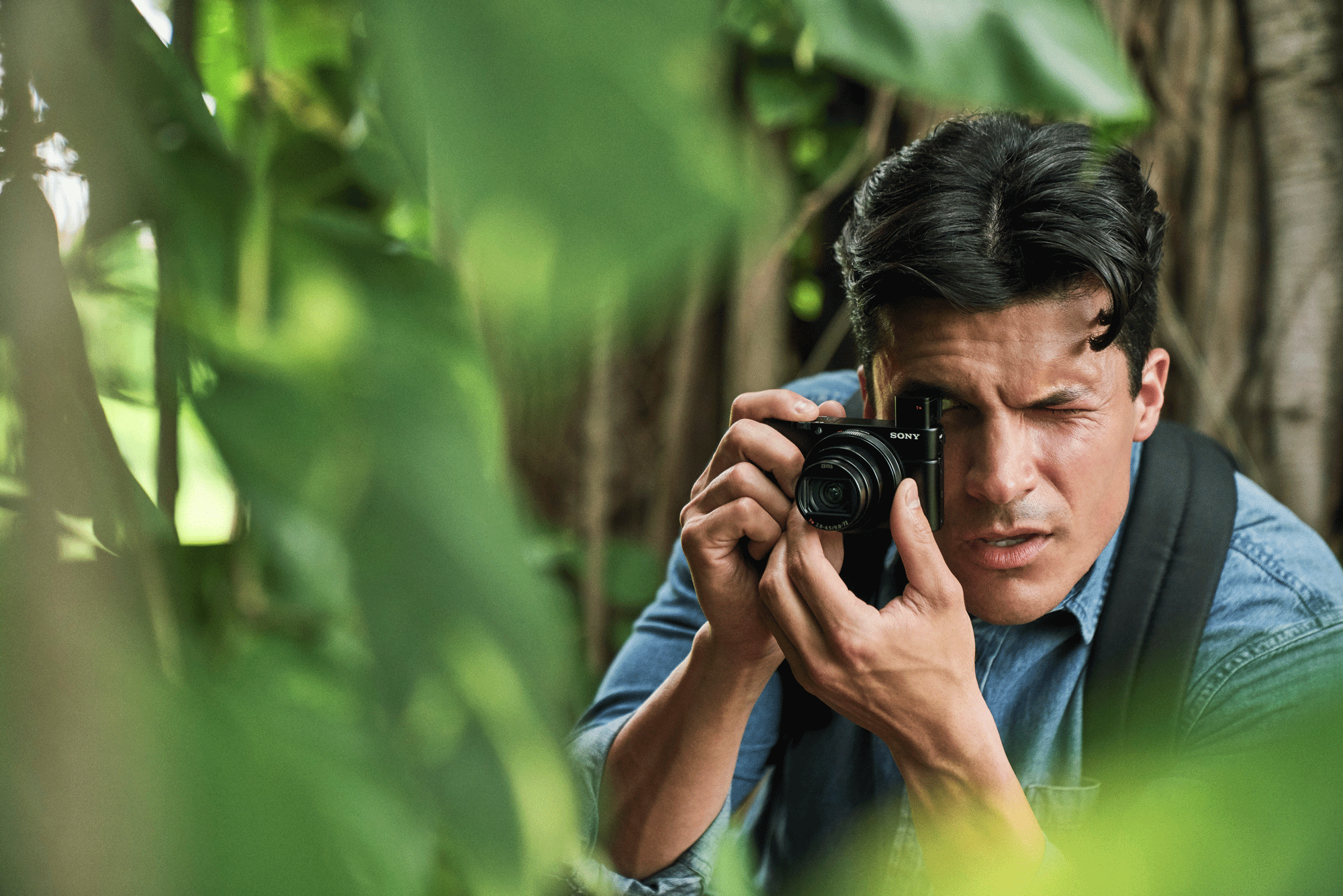 A man takes a photo with a camera. He is in a jungle-like scene, surrounded by green leaves and trees.