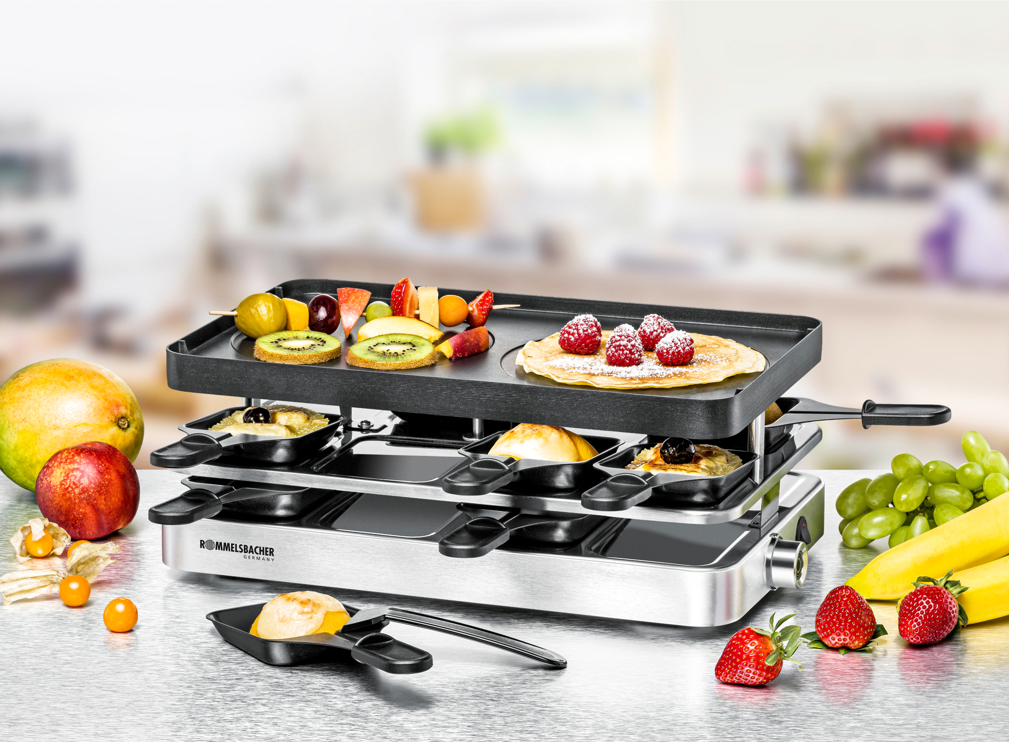 Raclette-Grill mit Essen. Obst, Pfannkuchen und Käse in Pfannen. Küchenhintergrund, verschiedene Früchte auf dem Tisch.