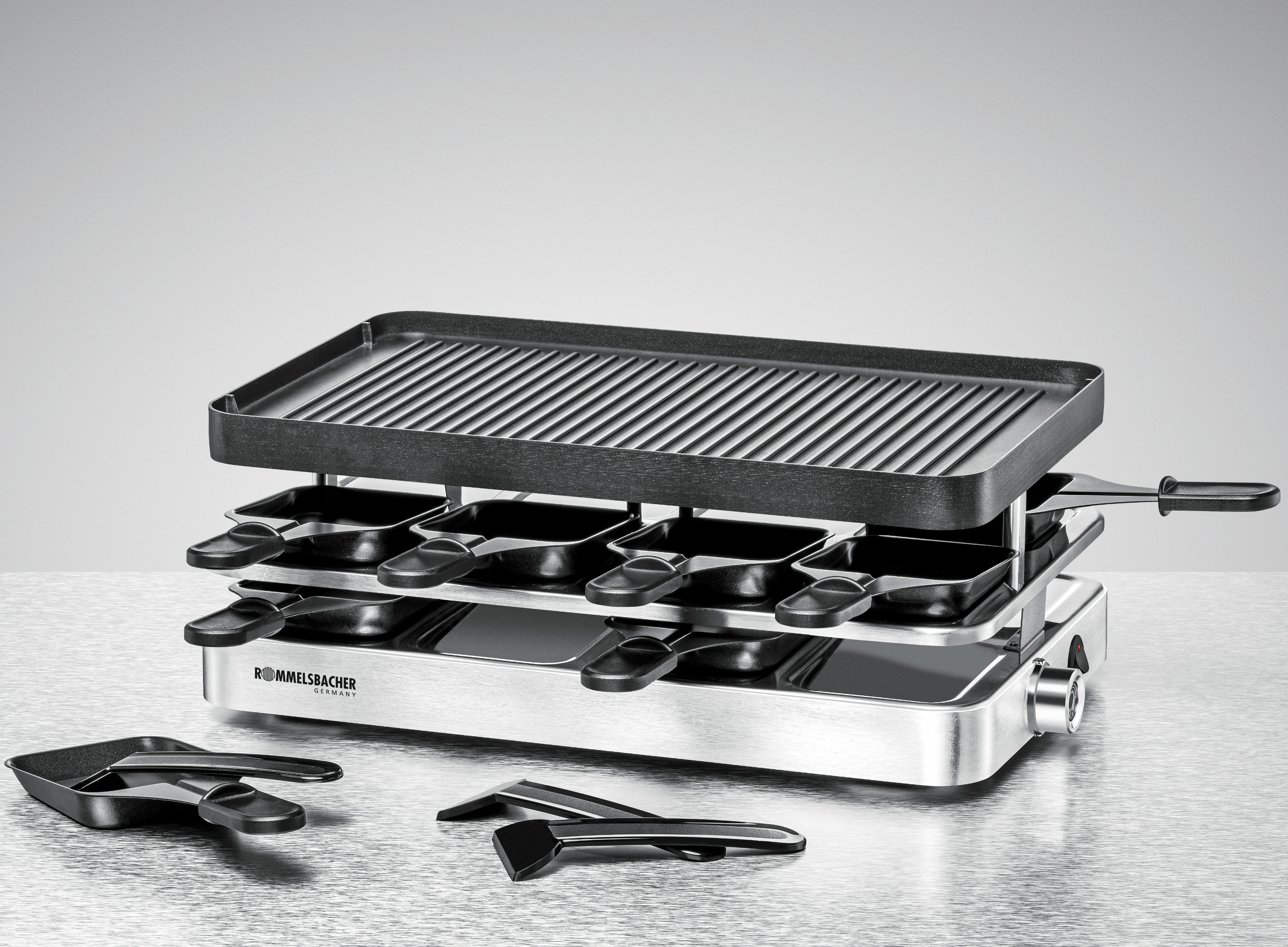 Raclette-Grill mit schwarzer Grillplatte und Käseschalen, auf Metalloberfläche.