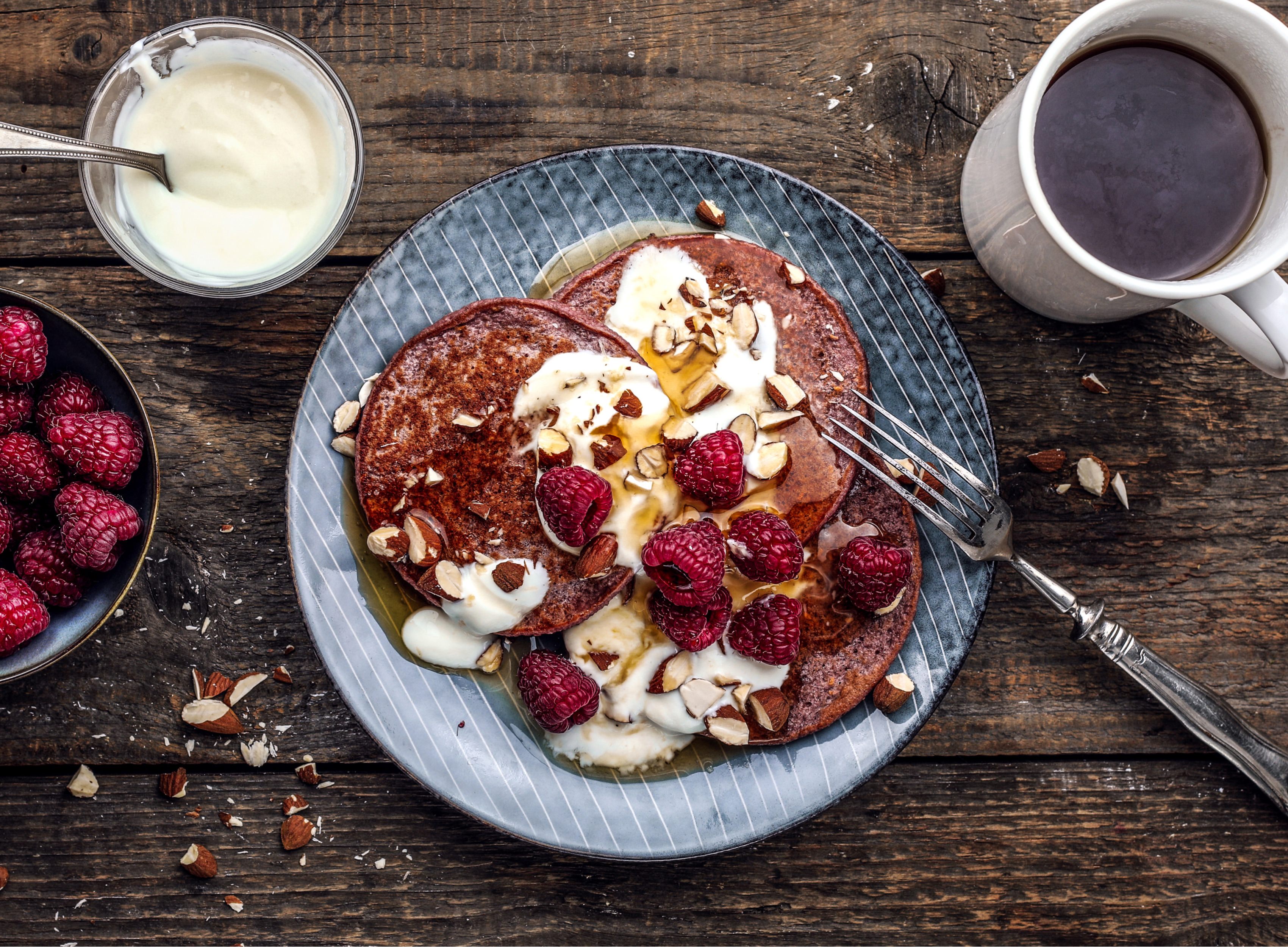 Pfannkuchen mit Himbeeren, Sahne und Mandeln auf einem Teller mit Kaffee und Joghurt.