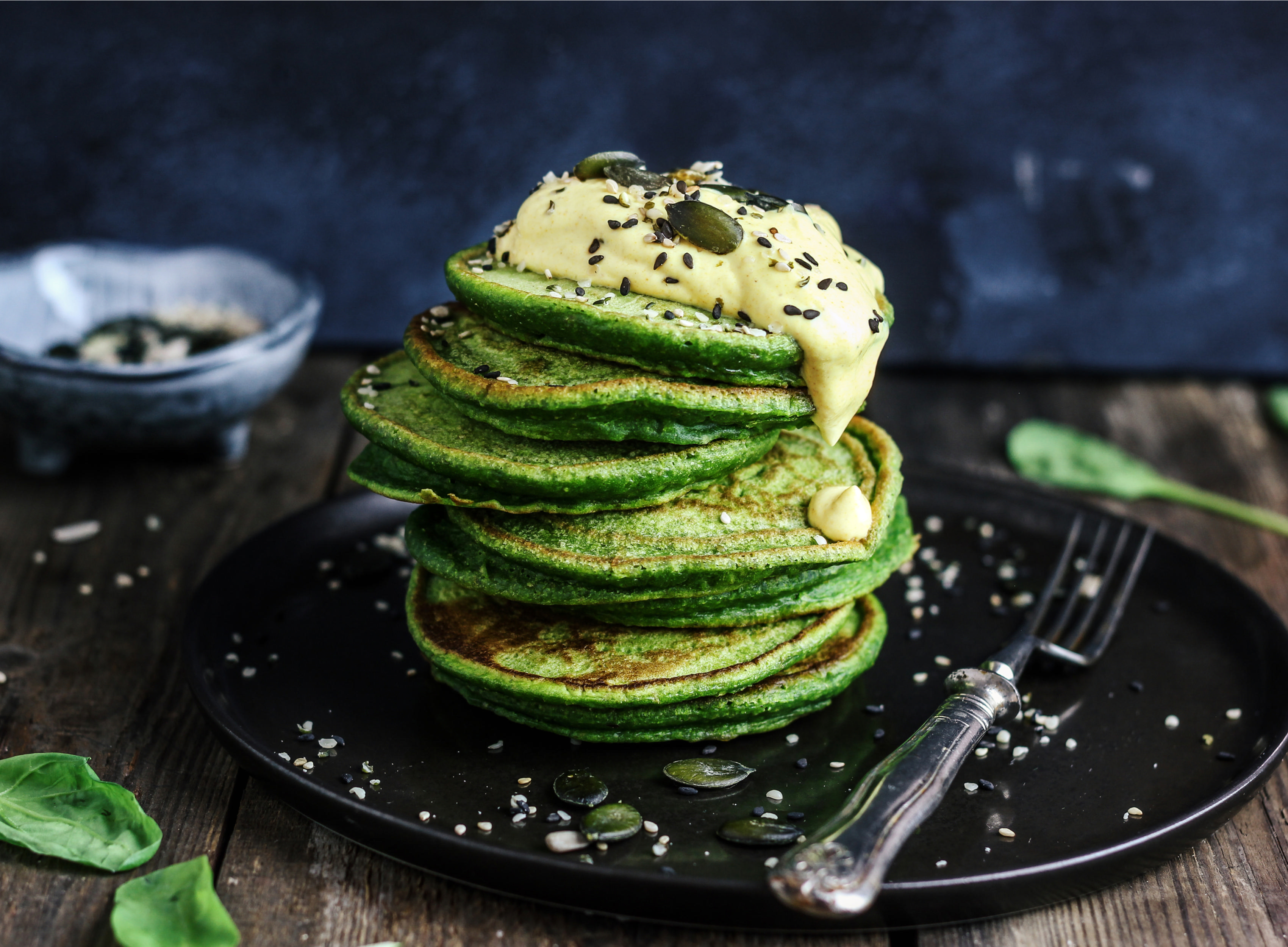 Grüne Pfannkuchen mit gelber Creme und Samen auf einem schwarzen Teller und einem Holztisch.
