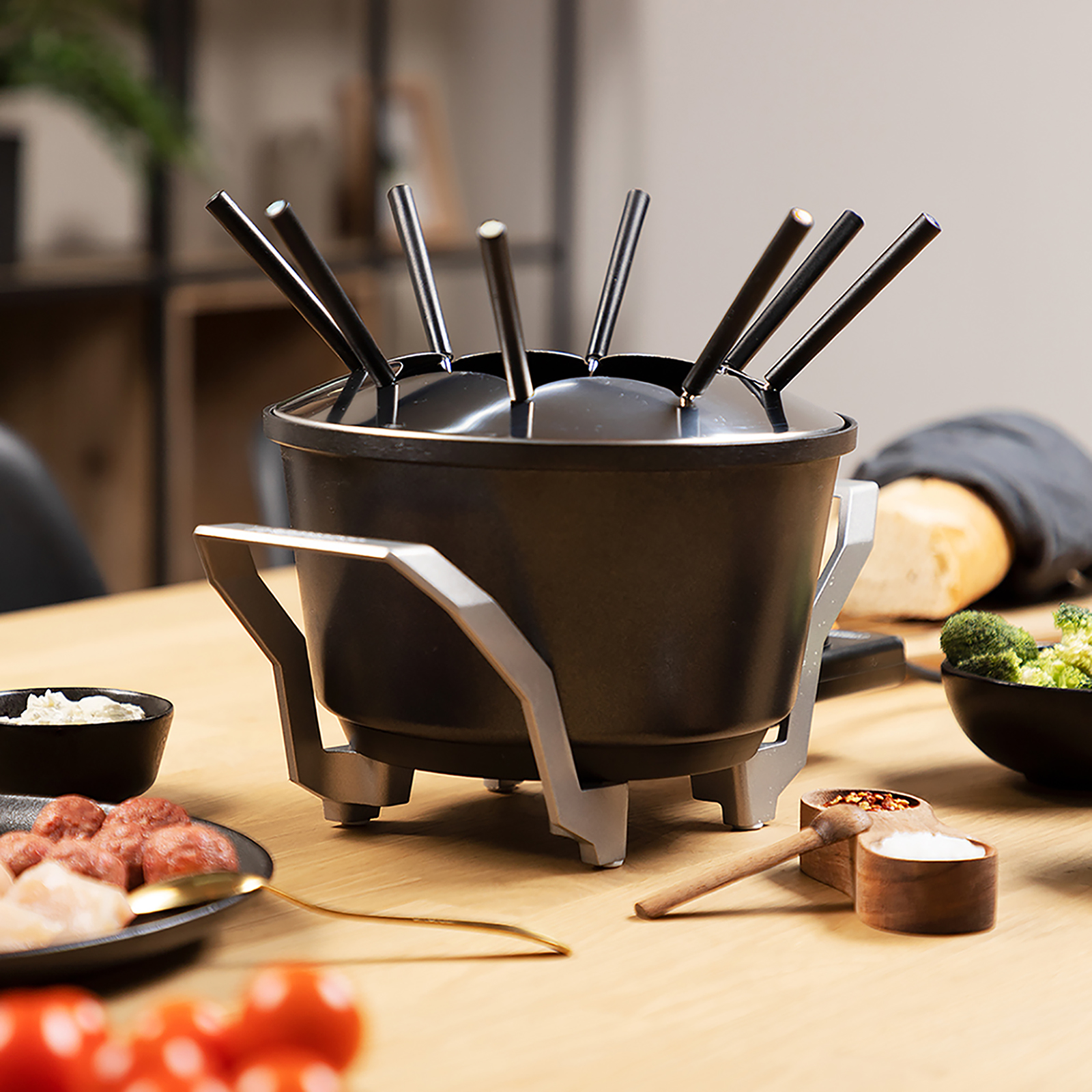 Fondue set on a wooden table. Ingredients and utensils are placed around it.