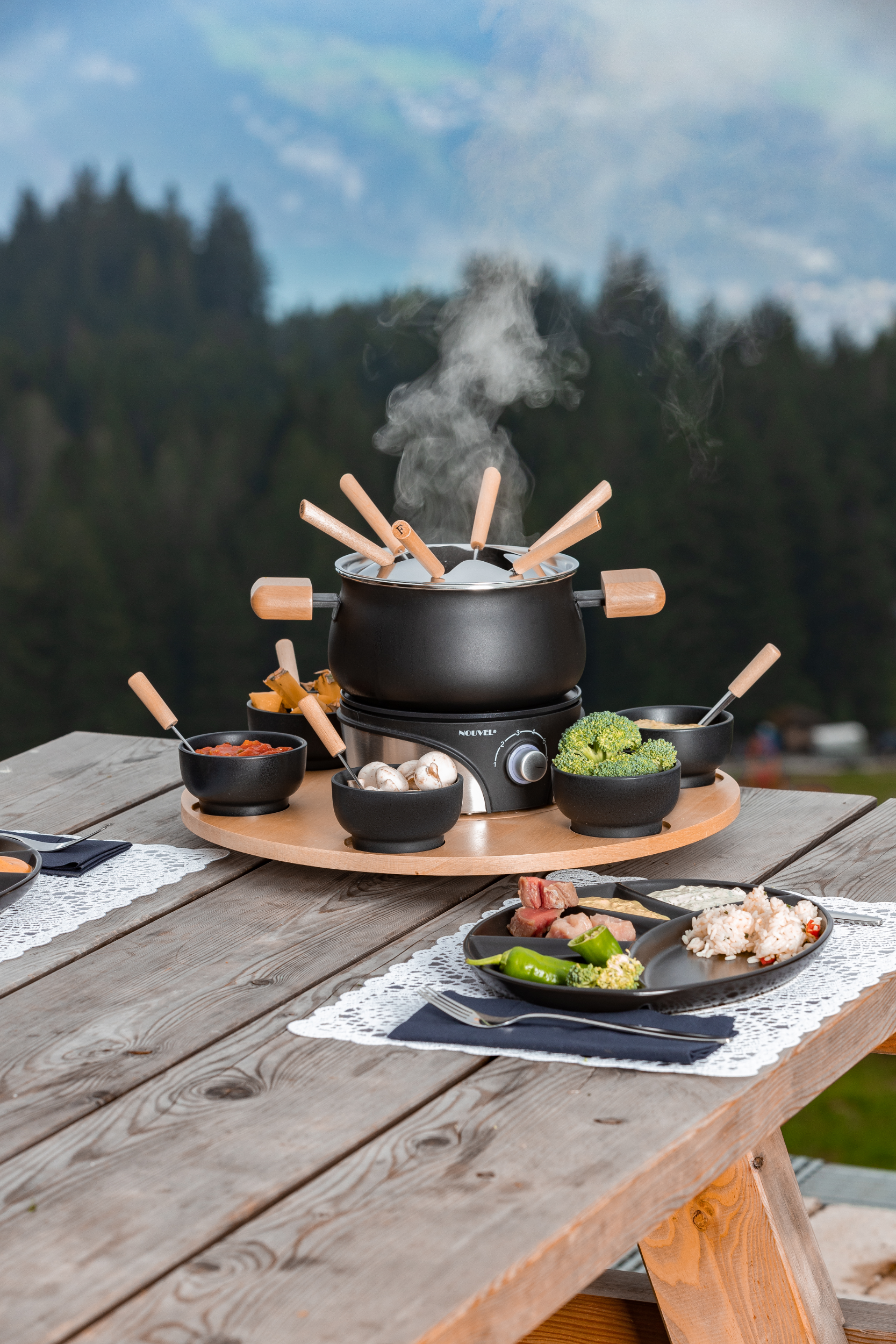 Set per fonduta su un tavolo di legno, con cibo intorno. Sfondo montano panoramico in lontananza.
