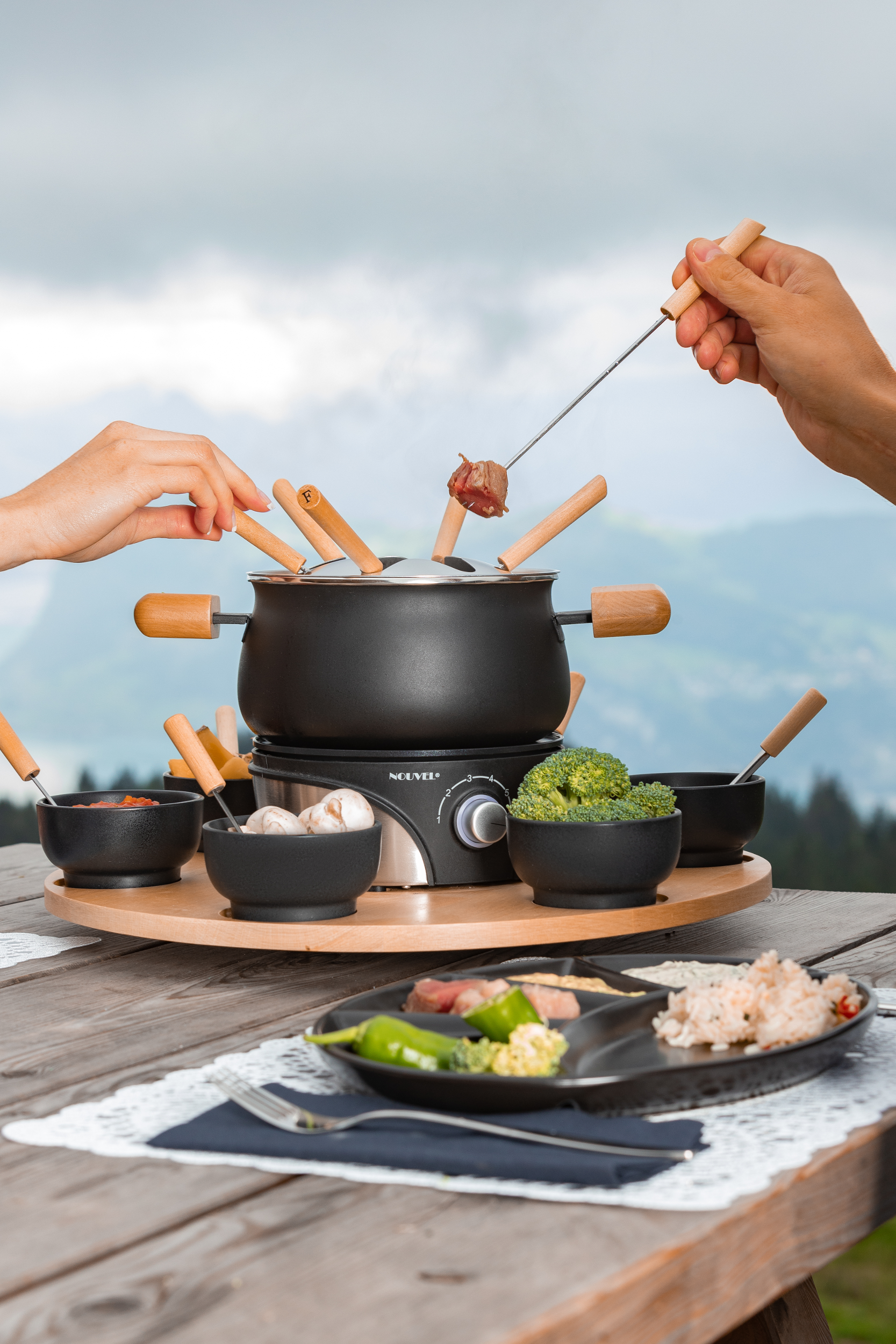 Set per fonduta su un tavolo di legno con cibo, mani che usano forchette per intingere.