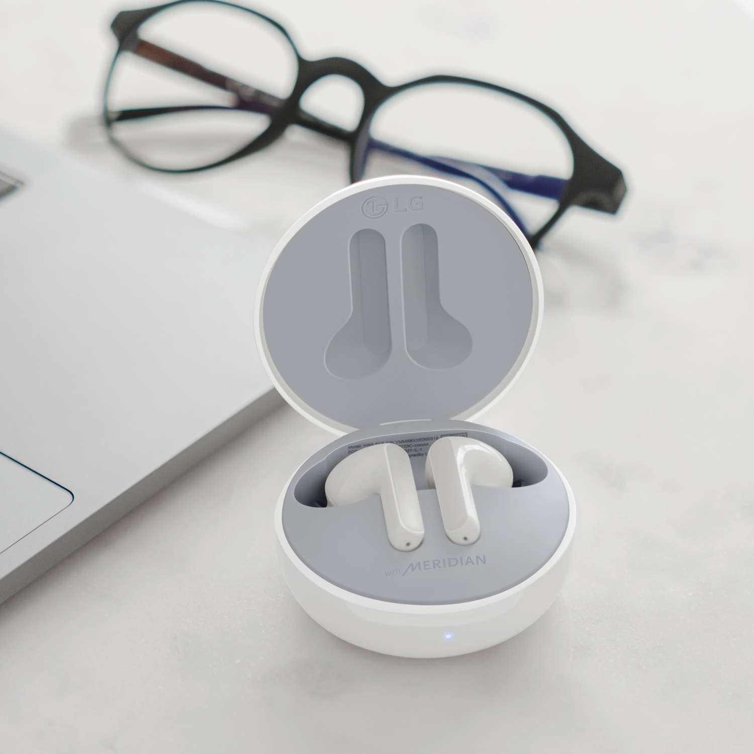 Auriculares inalámbricos LG blancos en un estuche de carga abierto, con gafas y portátil al fondo.