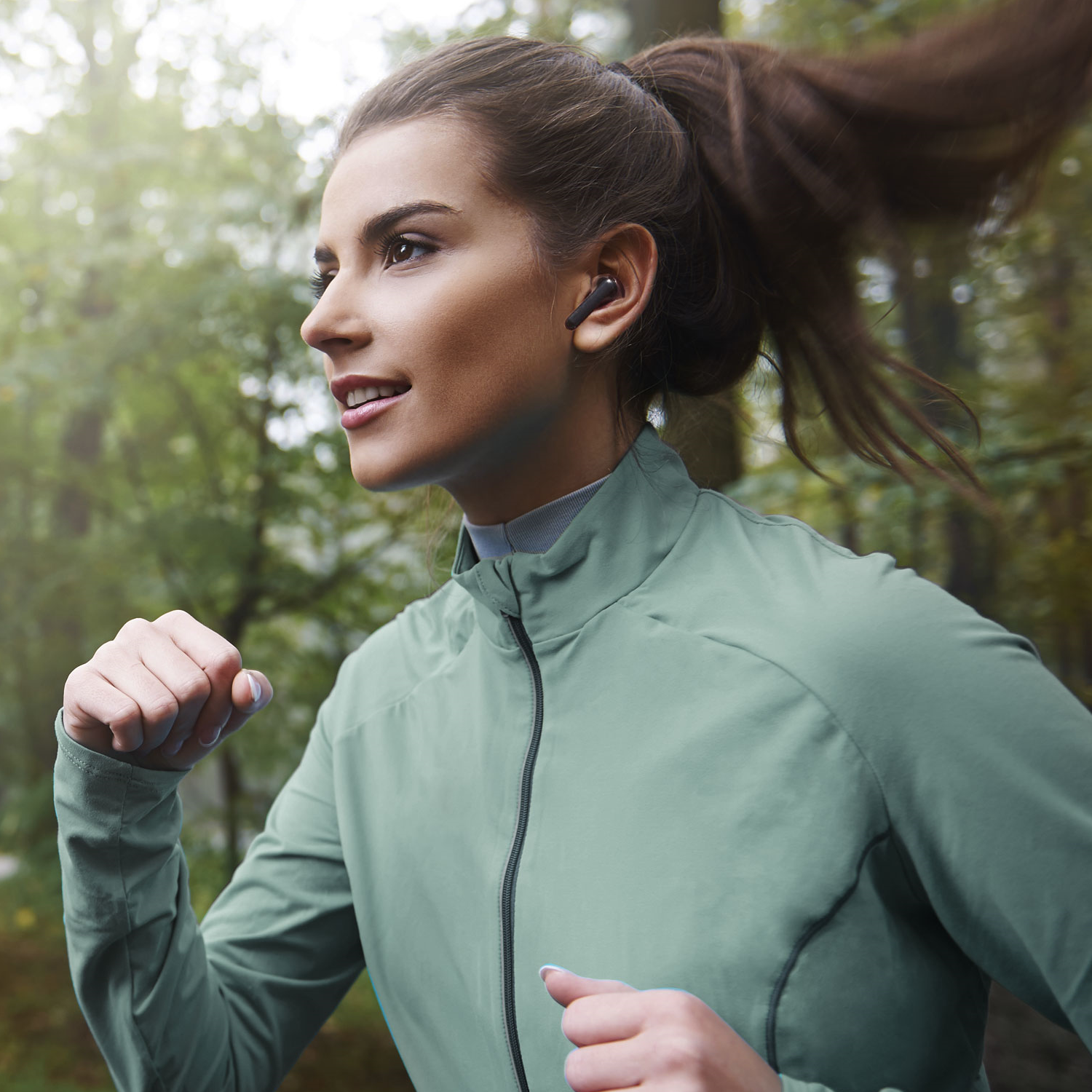 Una mujer corriendo por un bosque con una chaqueta verde y auriculares. Ella está sonriendo.
