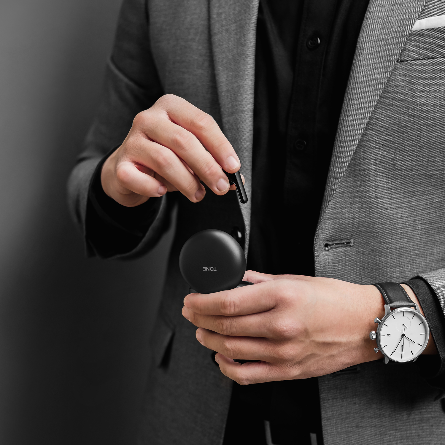 Un hombre con traje sostiene un estuche de carga negro y un auricular. Un reloj está en su muñeca.