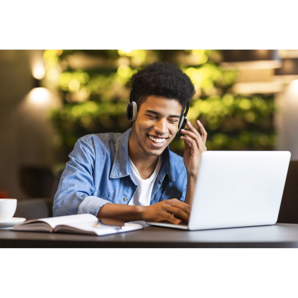 Hombre negro sonriente con auriculares usando una laptop. Cuaderno abierto, cafetería.