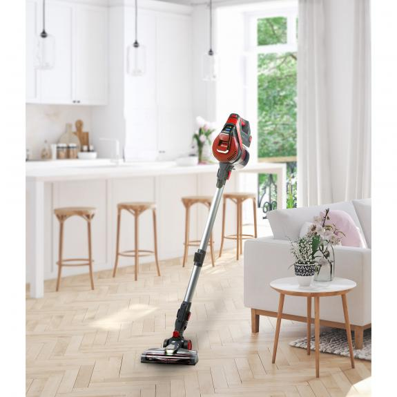 Un aspirateur rouge et noir est sur un sol en bois. Cuisine et salon blancs en arrière-plan.