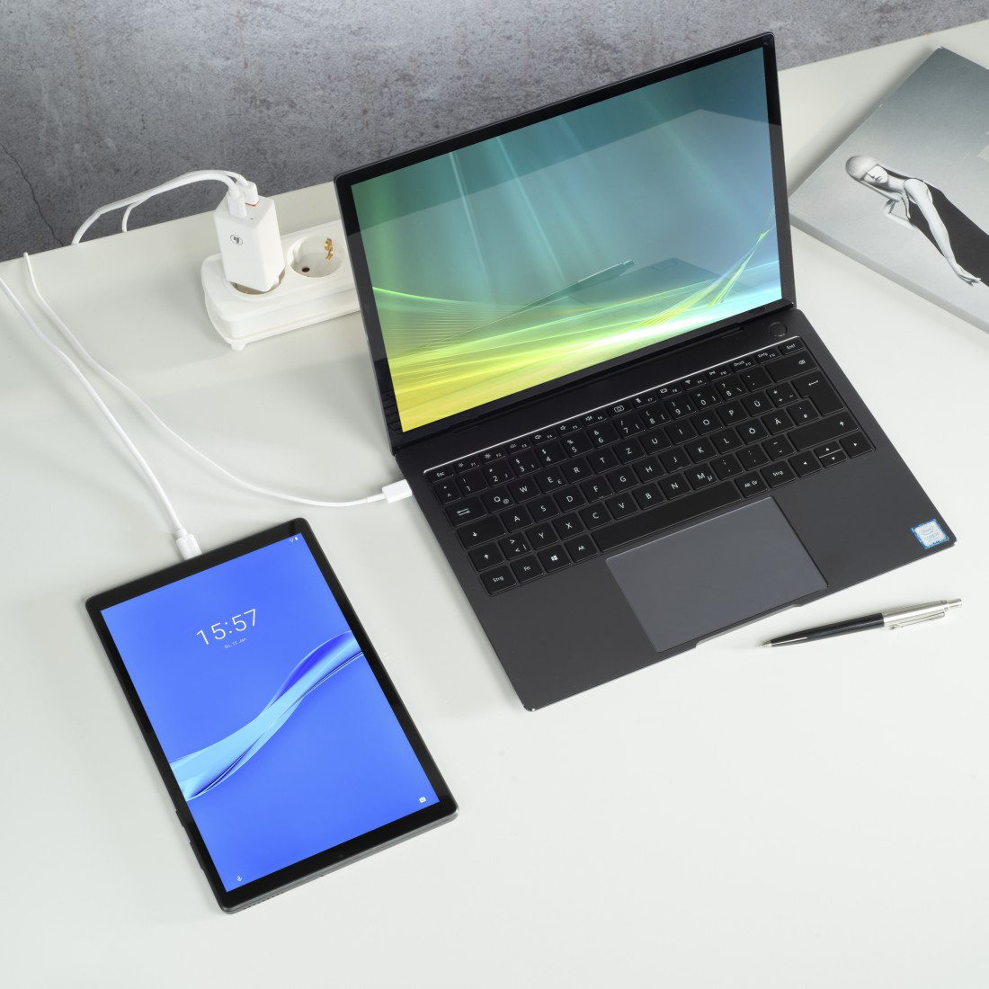Tablet und Laptop verbunden, laden auf einem weißen Schreibtisch. Steckdose und Magazin.