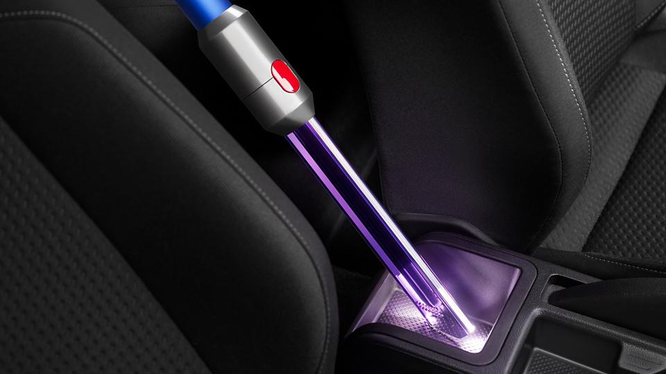 Dyson Staubsauger reinigt einen Autoinnenraum. Die Düse ist lila, die Sitze sind schwarz.