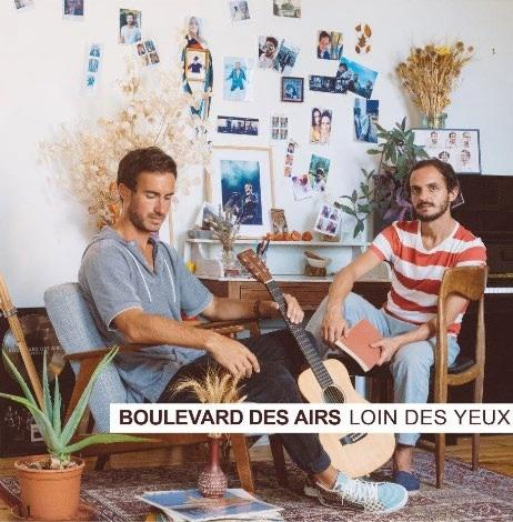 Deux hommes assis avec une guitare et un livre. Le mur a des photos et des plantes. Texte : Boulevard des Airs.