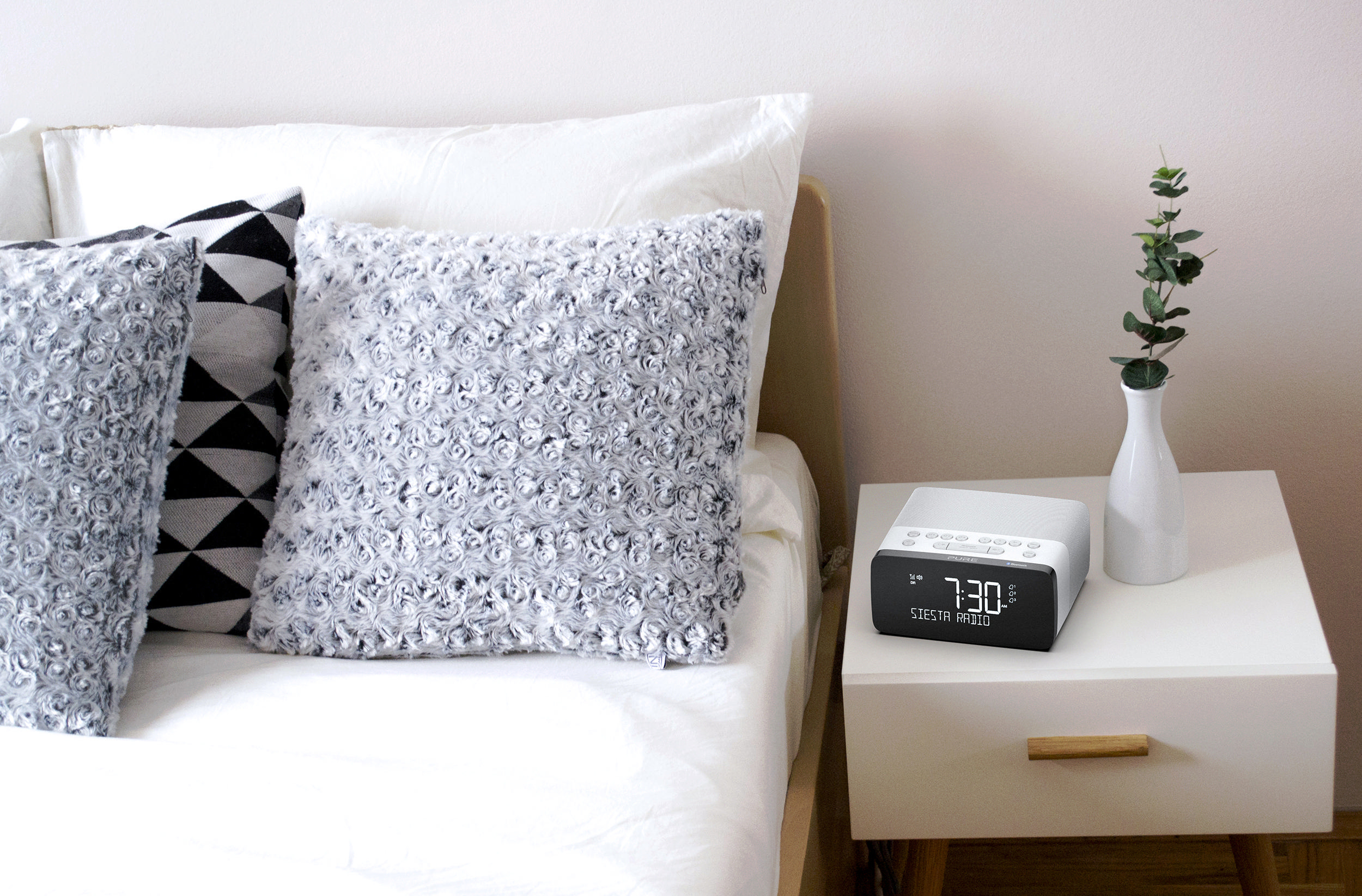 Bett mit Kissen und Nachttisch mit Uhr und Vase.
