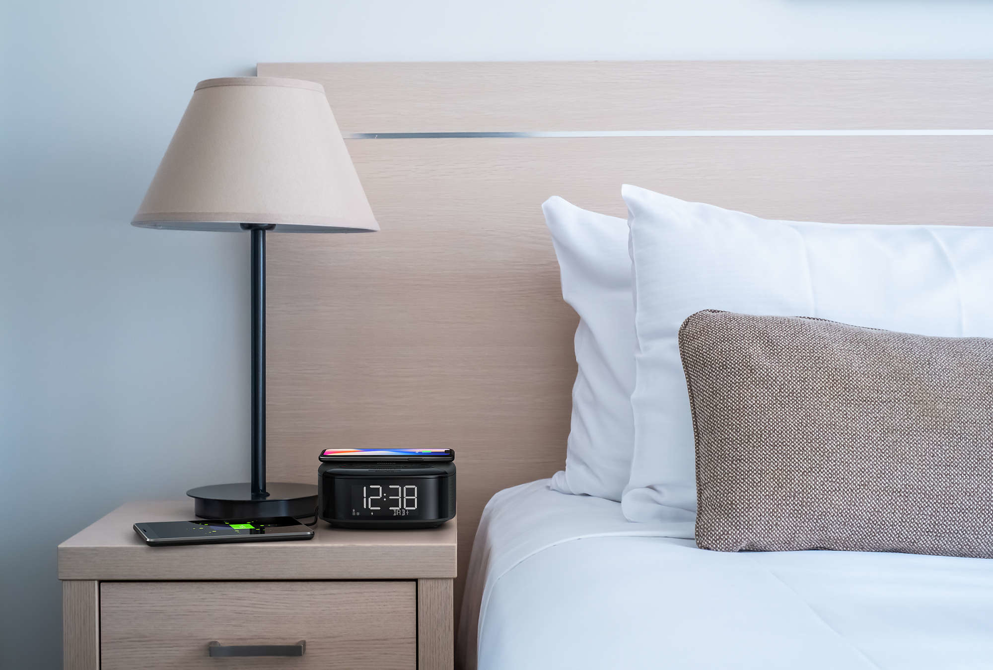 A nightstand with a lamp and a clock. A phone charges on the clock and another on the stand.