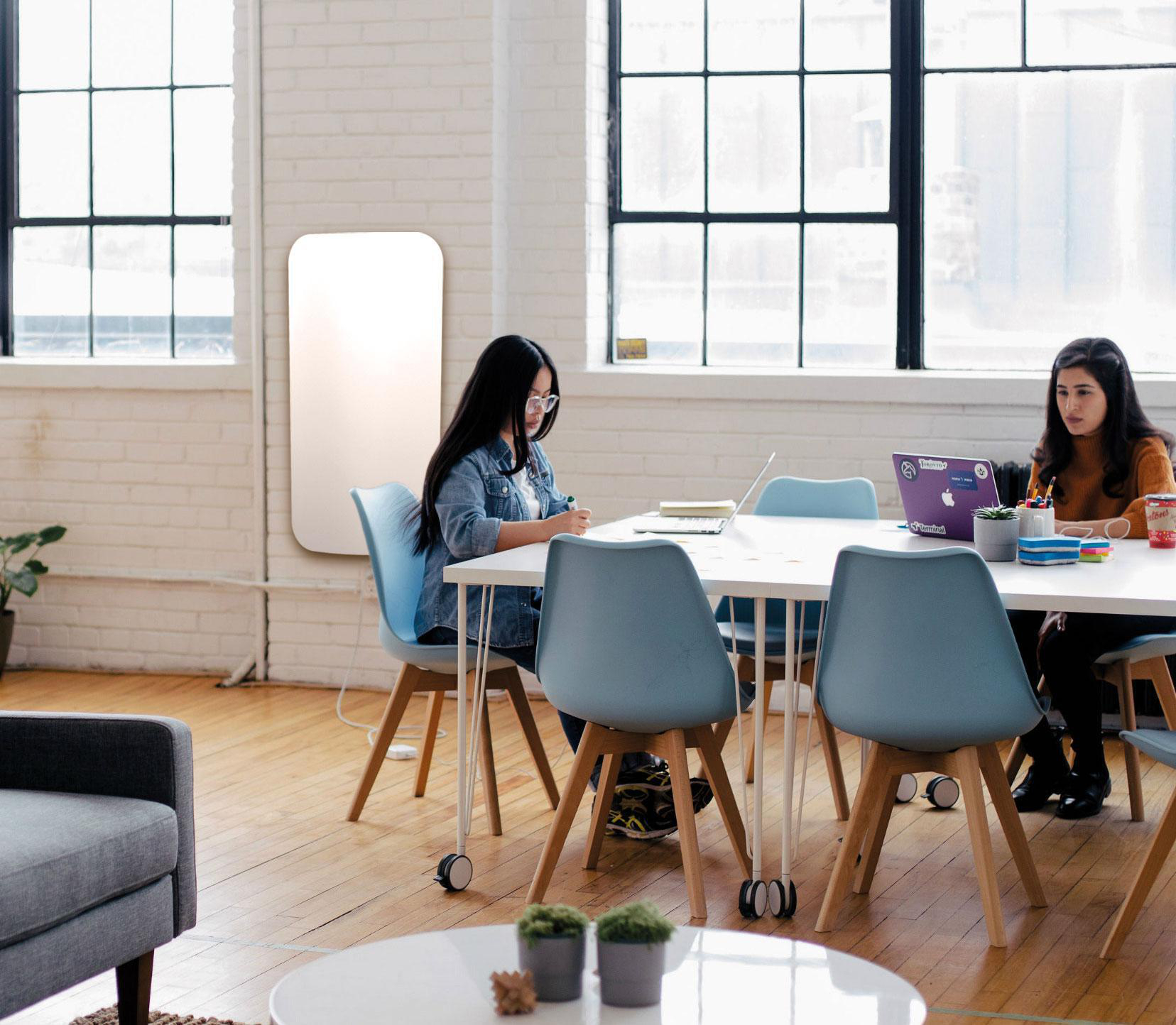 Zwei Frauen sitzen in einem Büro an einem Schreibtisch. Eine arbeitet an einem Laptop, die andere arbeitet.