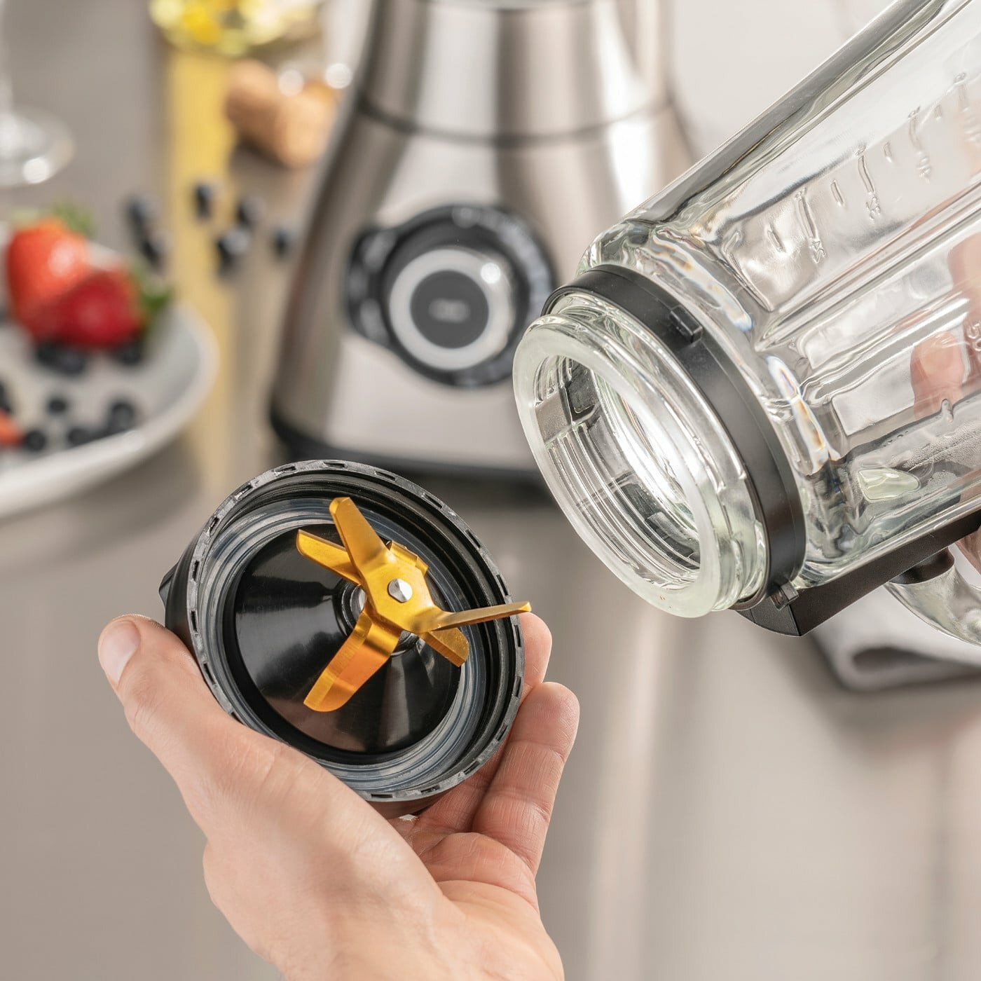 Eine Hand hält einen Mixer-Klingenaufsatz neben einem Glas, mit einem Mixer im Hintergrund.