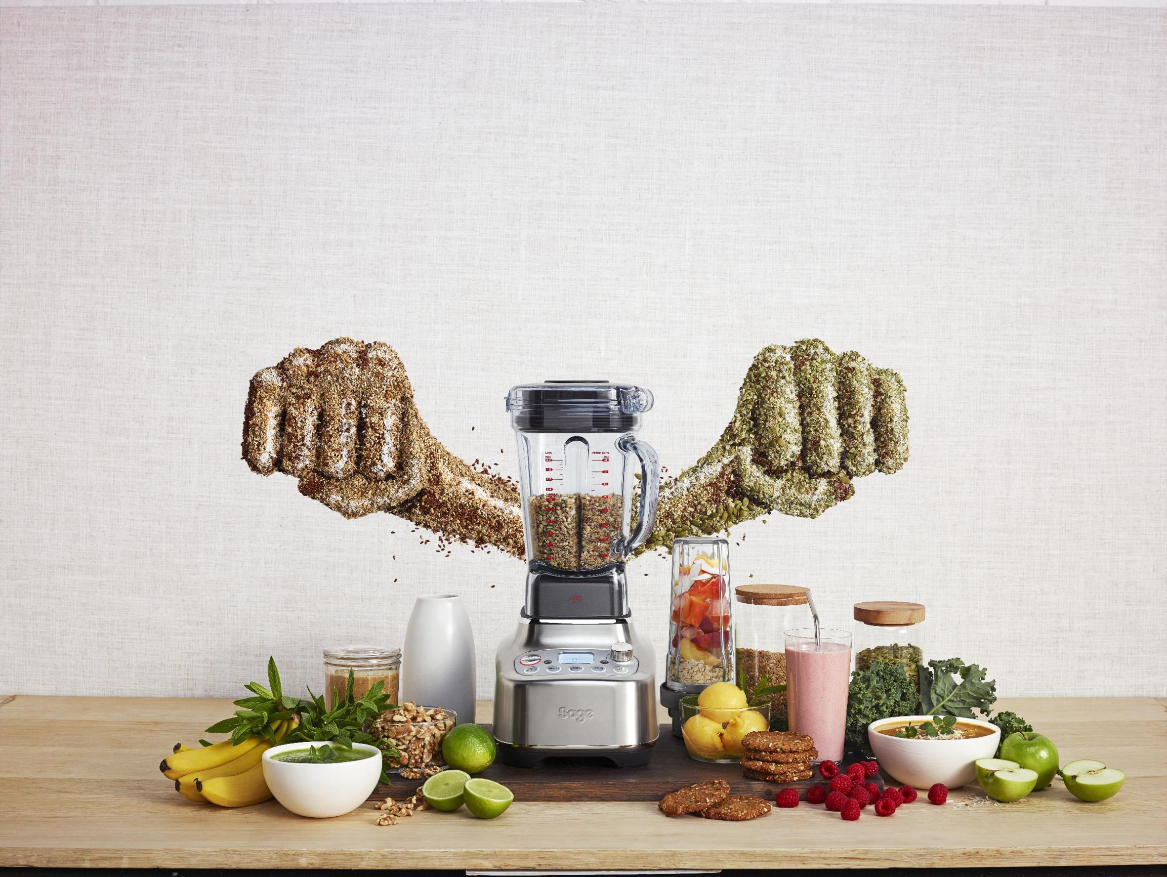 Mixer mit Früchten und Gemüse, mit zwei Fäusten aus Samen, auf einem Holztisch mit weißer Wand.
