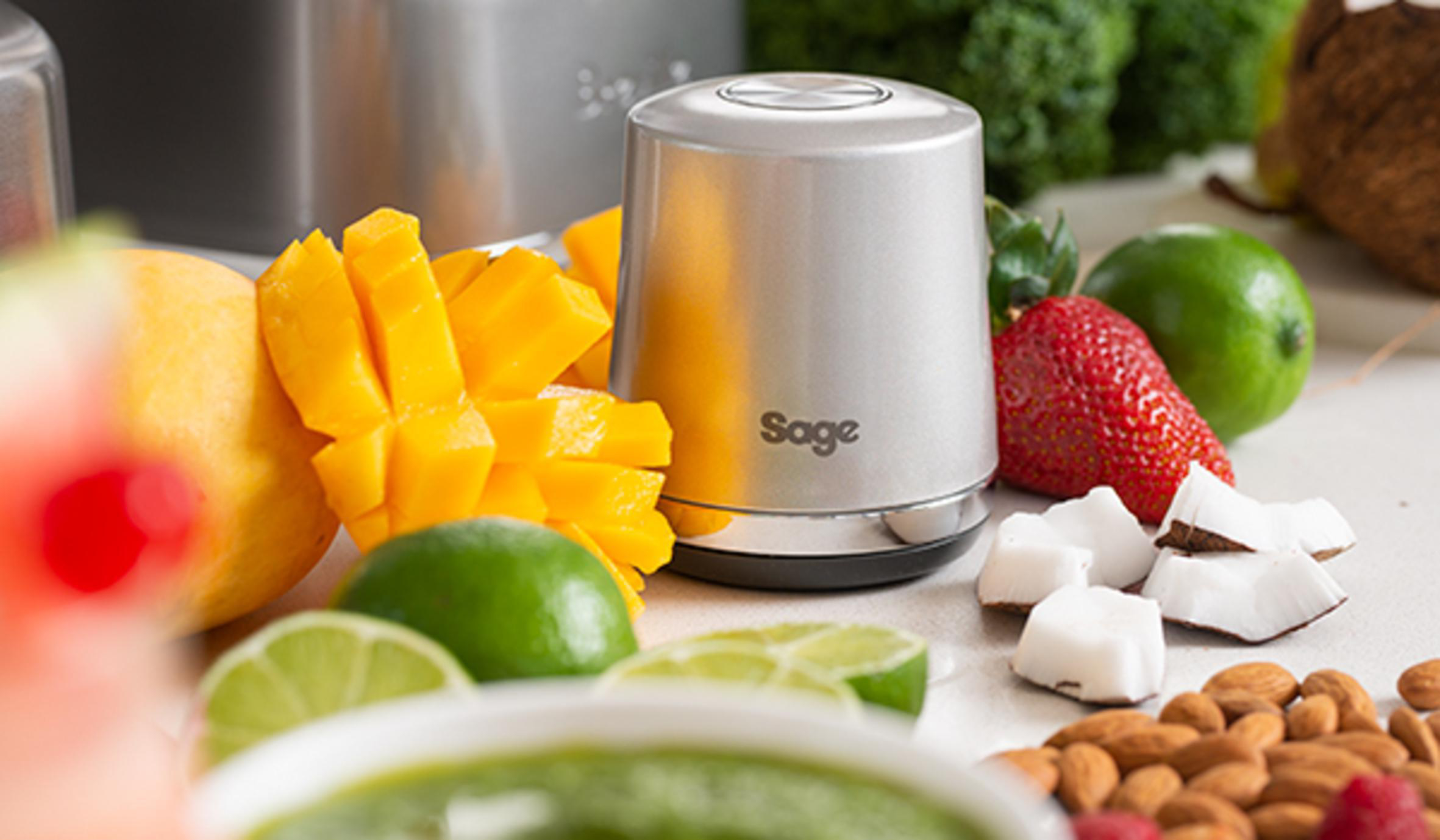 Silberner Sage-Foodprozessor mit Lebensmitteln drumherum. Mango, Limette, Kokosnuss und andere Früchte.