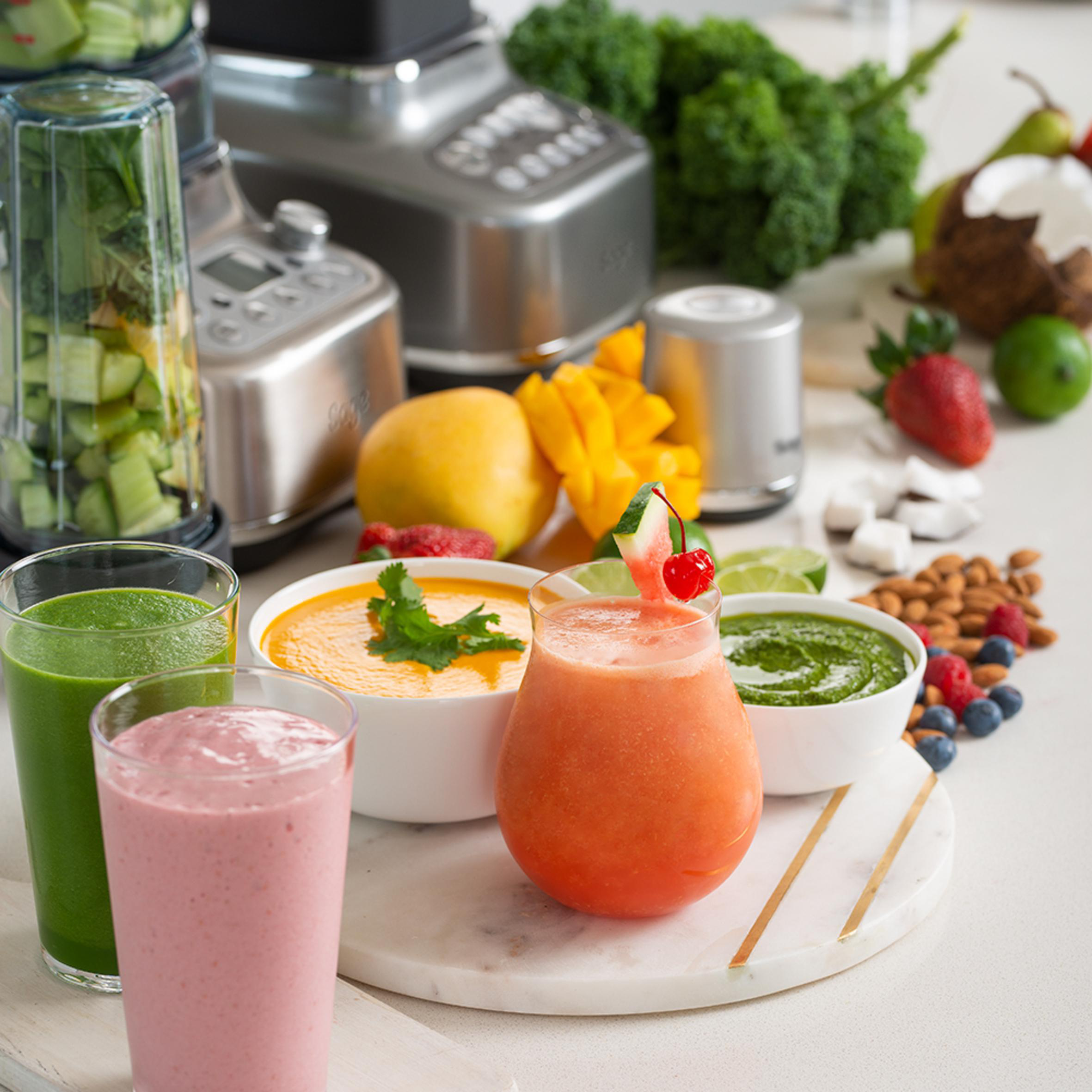 Verschiedene bunte Smoothies, frische Früchte und ein Mixer im Hintergrund.