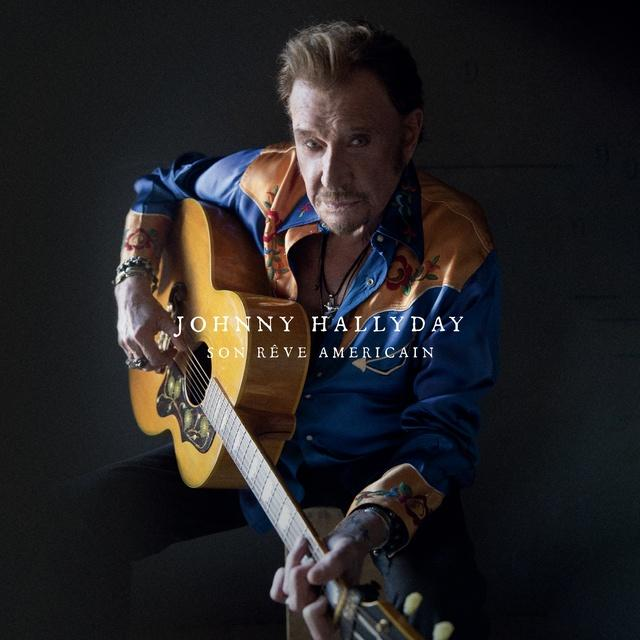 Johnny Hallyday holding a guitar, looking at the camera, with blue and orange shirt. Background is dark.