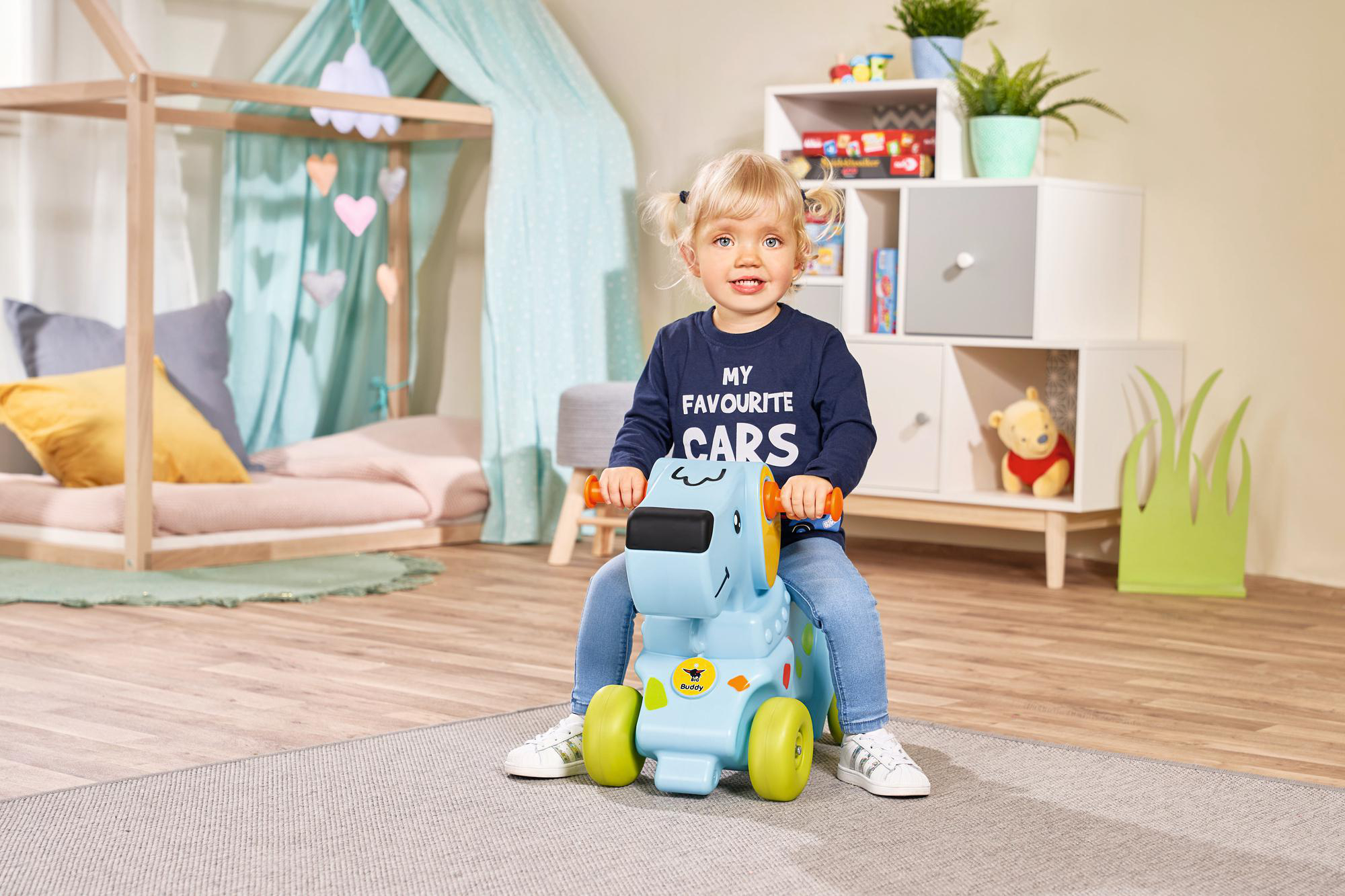 Ein Kleinkind auf einem blauen Spielzeugauto lächelnd, in einem Raum mit Spielzeug und Deko.