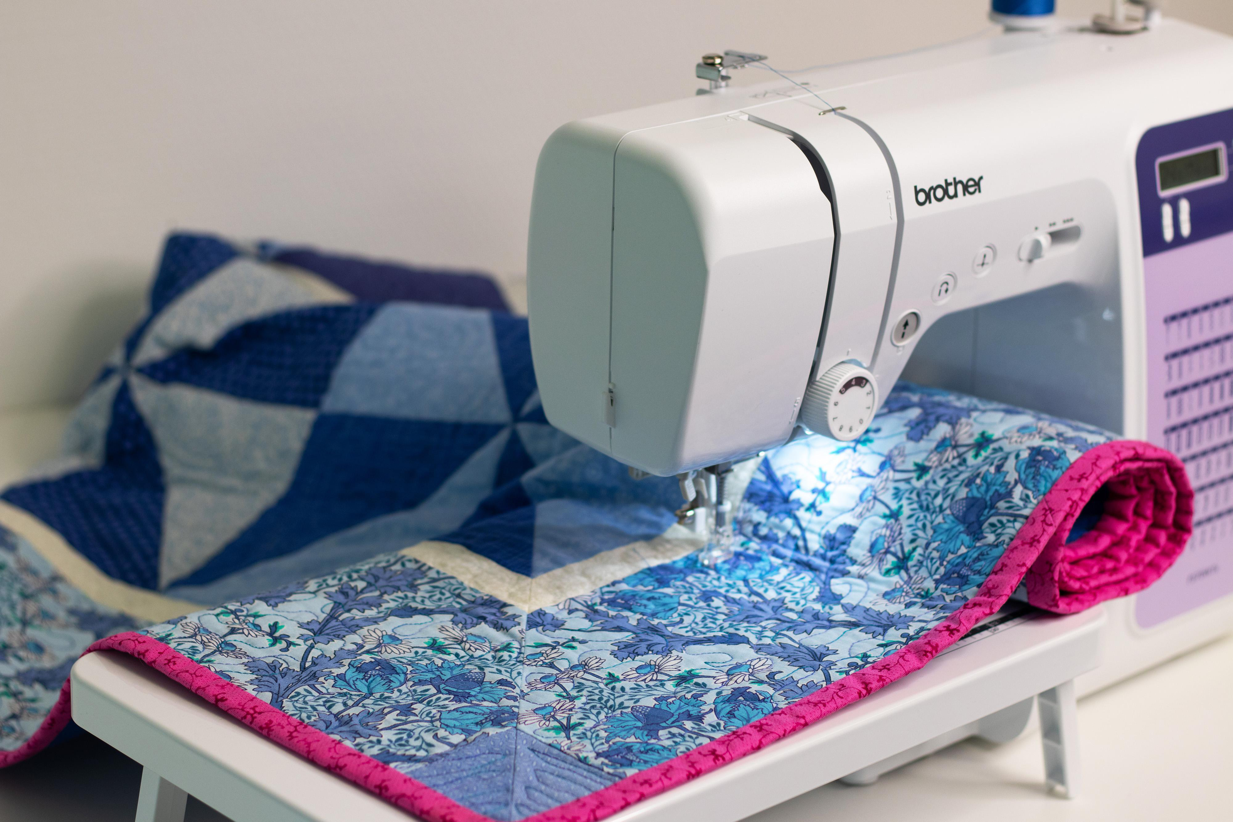 Eine weiße Brother-Nähmaschine näht an einem blau-gemusterten Quilting-Stoff mit rosa Rand.