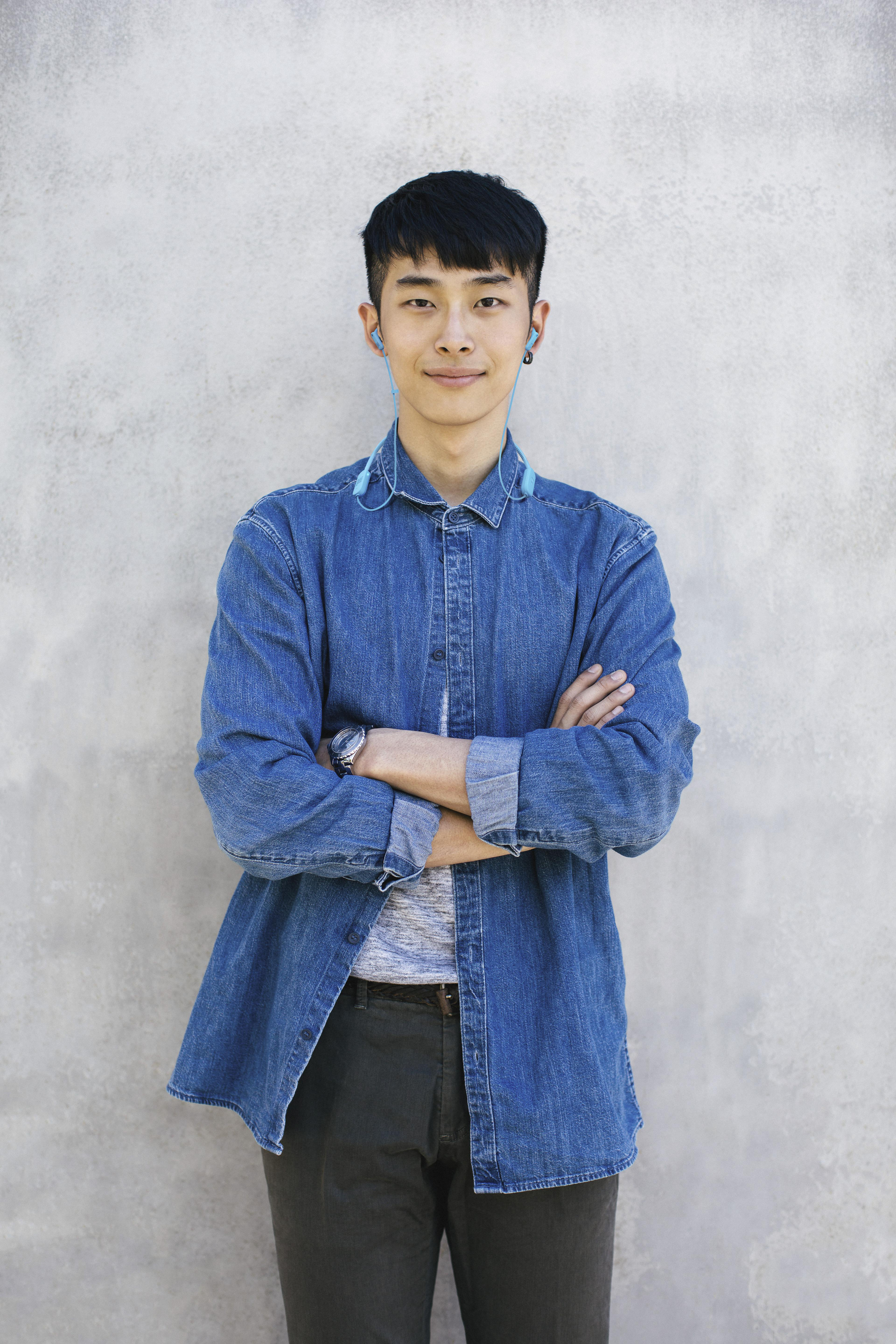 Junger asiatischer Mann mit Jeanshemd und verschränkten Armen, blaue Ohrhörer. Heller Hintergrund.