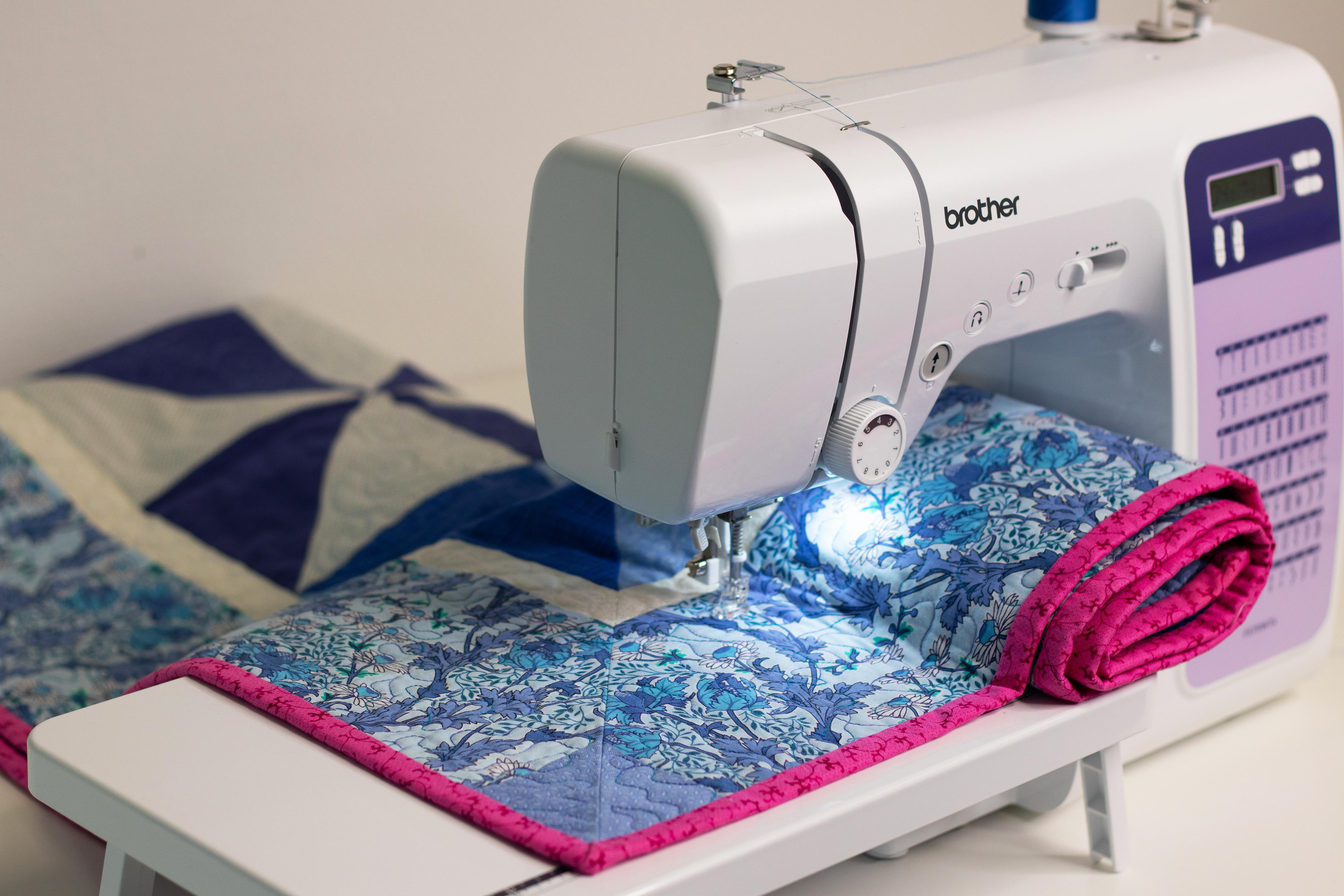 Eine weiße Brother-Nähmaschine näht einen blauen Blumenquilt mit rosa Einfassung.