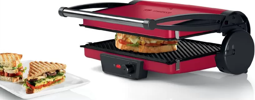 Presse à sandwich rouge avec un sandwich à l'intérieur et deux sandwichs finis sur une assiette blanche, prise de studio.