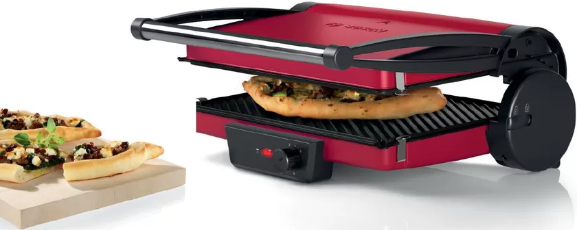 Une presse à sandwich rouge avec un sandwich à l'intérieur et deux bateaux de pain sur une planche en bois.