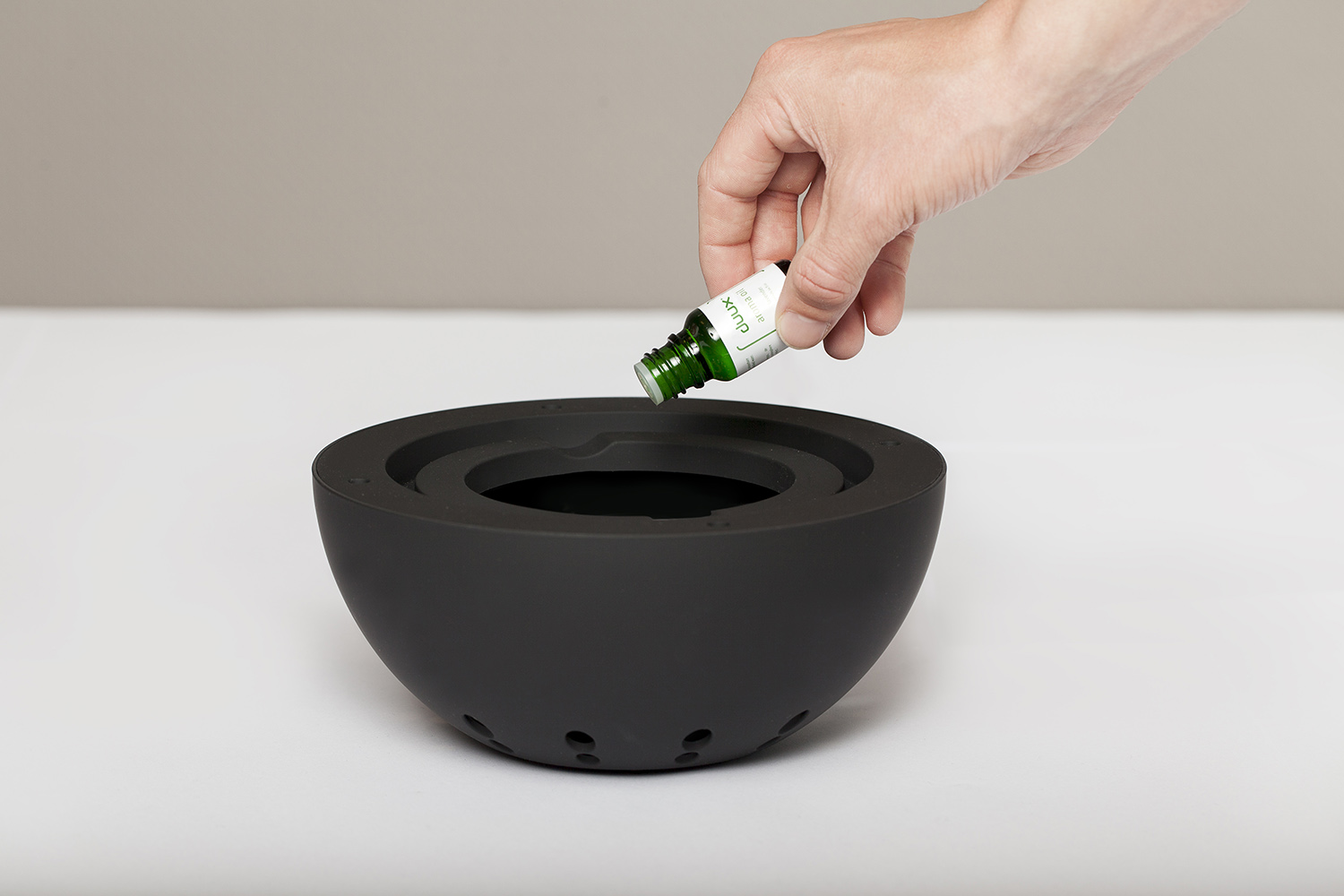A hand pouring green liquid from a bottle into a black aromatherapy diffuser bowl.