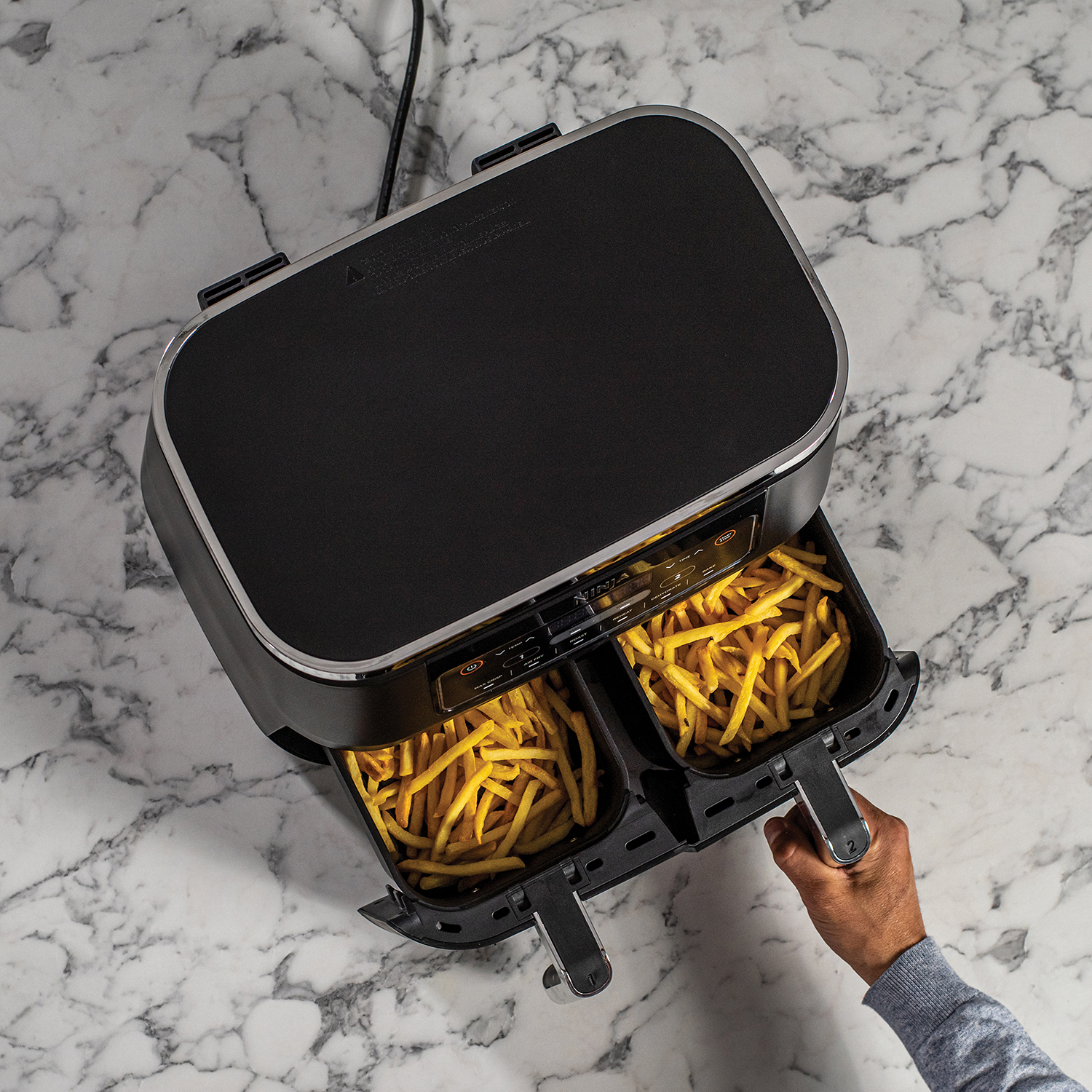Een airfryer met twee compartimenten gevuld met friet. Een hand houdt hem vast.