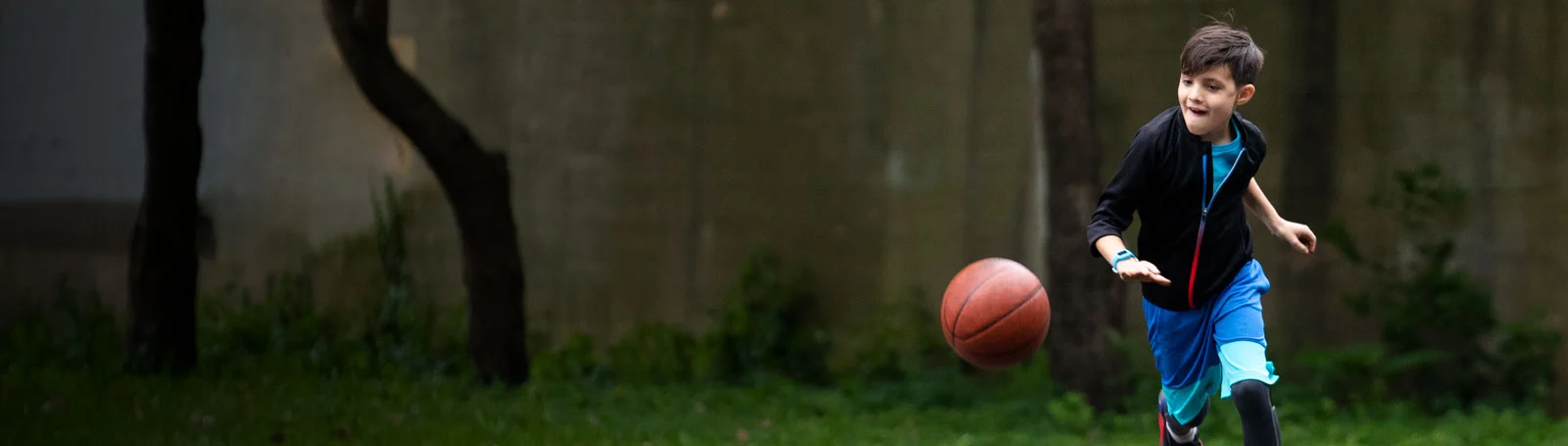 Garçon en tenue de sport dribble un ballon de basket. Herbe verte et arbres en arrière-plan.