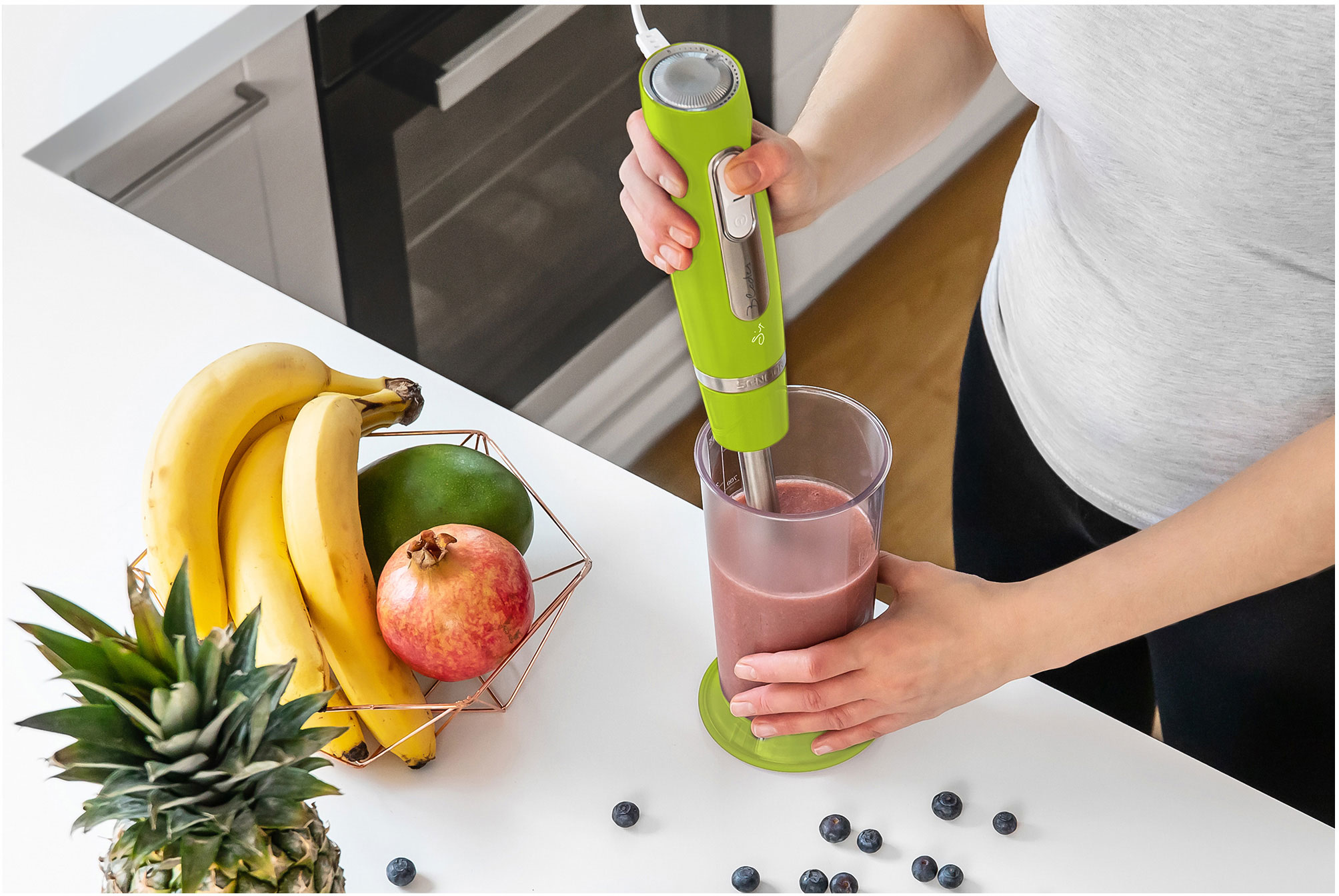 Egy nő zöld botmixerrel turmixot készít. Gyümölcsök egy kosárban, fehér felületen.